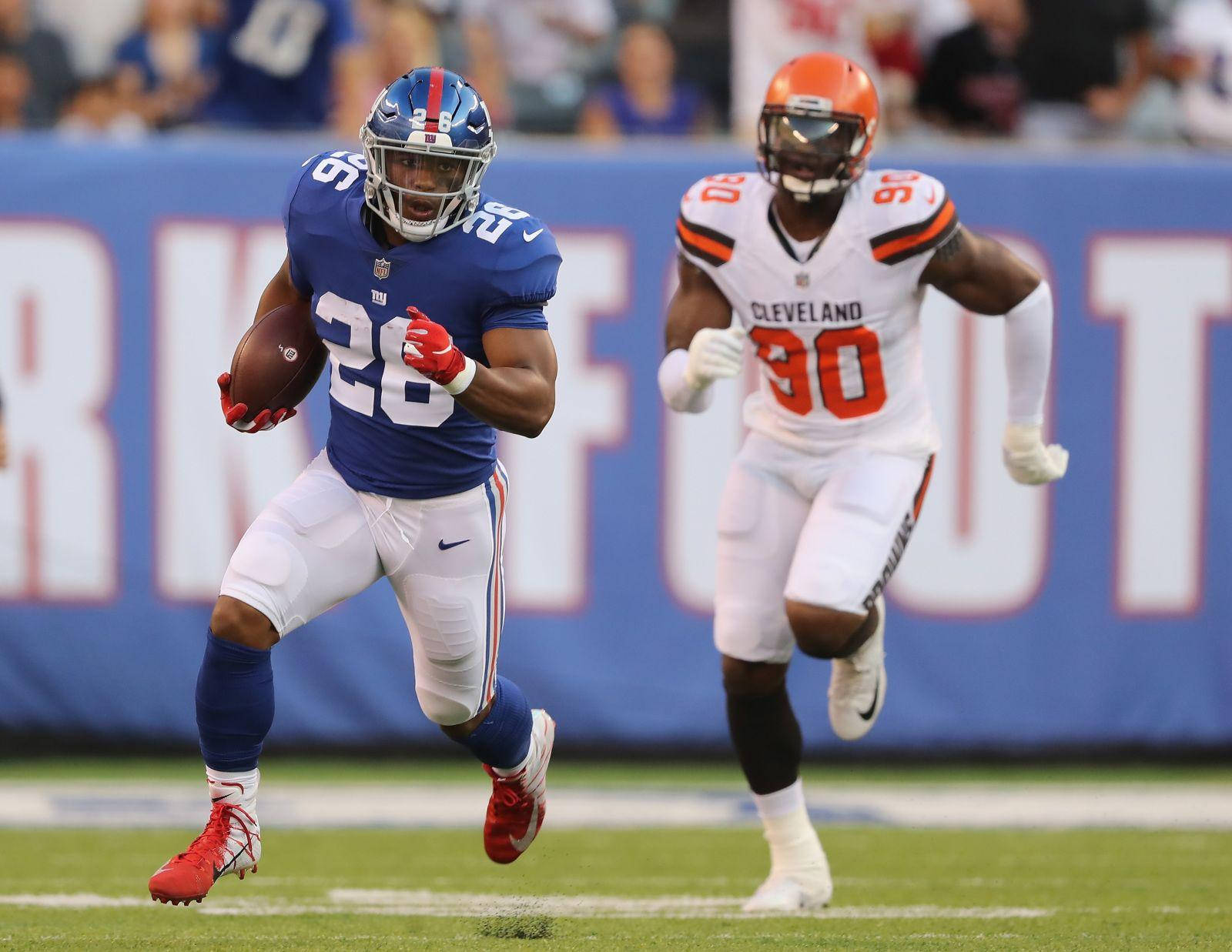 Saquon Barkley 桌布 壁紙 — 一位身穿藍色球衣的紐約巨人隊球員帶著美式足球奔跑，而一位身穿白色球衣的克里夫蘭布朗隊防守球員在綠色球場上追趕他。