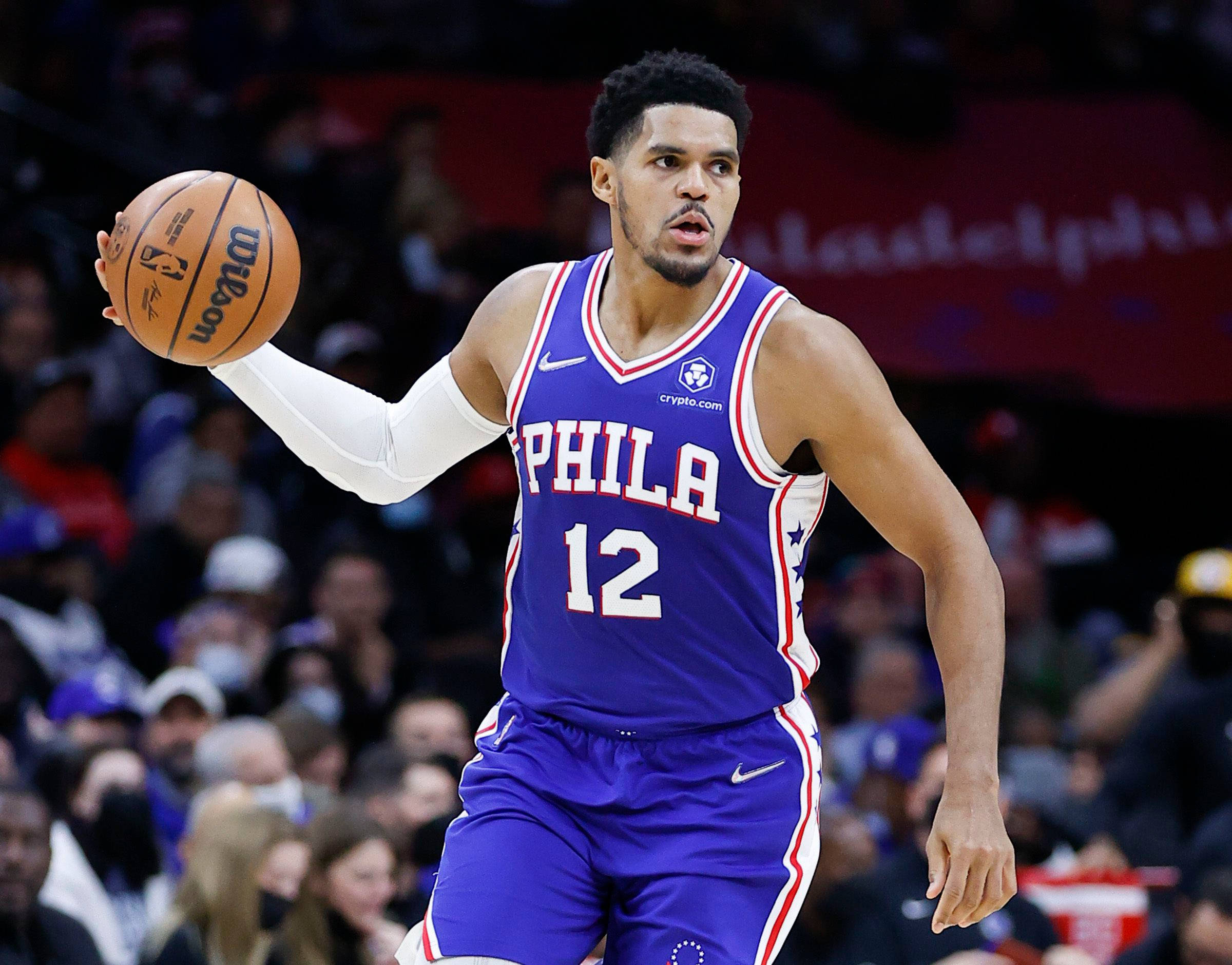 NBA 桌布 壁紙 — 一位身穿藍色球衣的費城76人籃球運動員在激烈的比賽中運球，背景是模糊的人群。