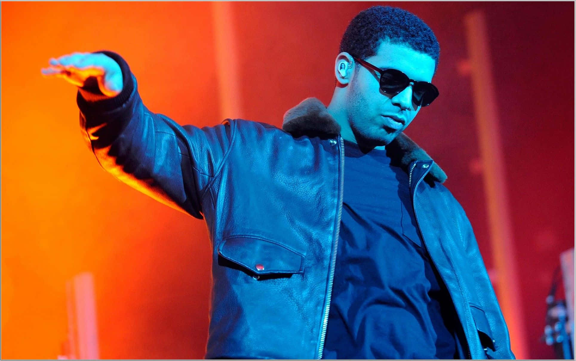 Drake 桌布 壁紙 — 一位身穿藍色皮夾克和太陽眼鏡的表演者在充滿活力的橙色和藍色舞台燈光下自信地做手勢。
