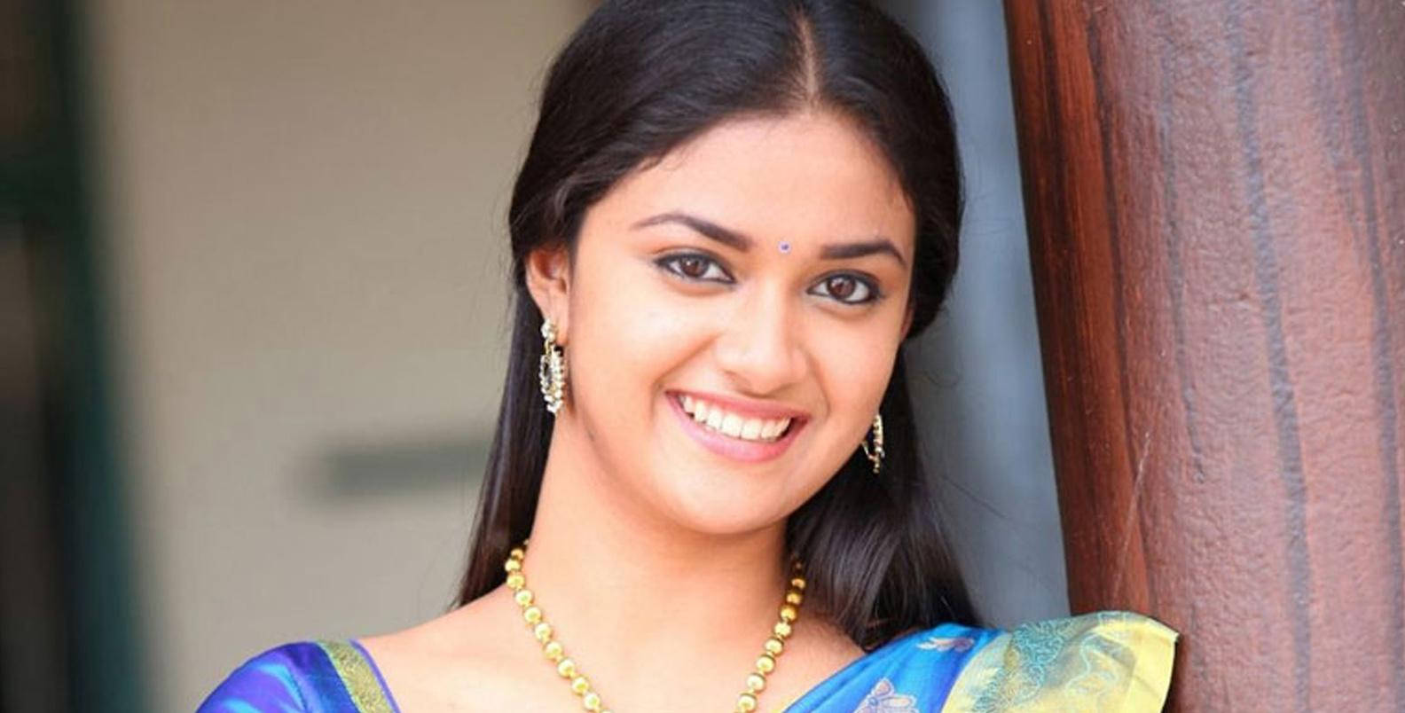 Keerthi Suresh HD 桌布 壁紙 — 一位身穿藍色紗麗的耀眼女性靠著木柱，露出溫暖的笑容。