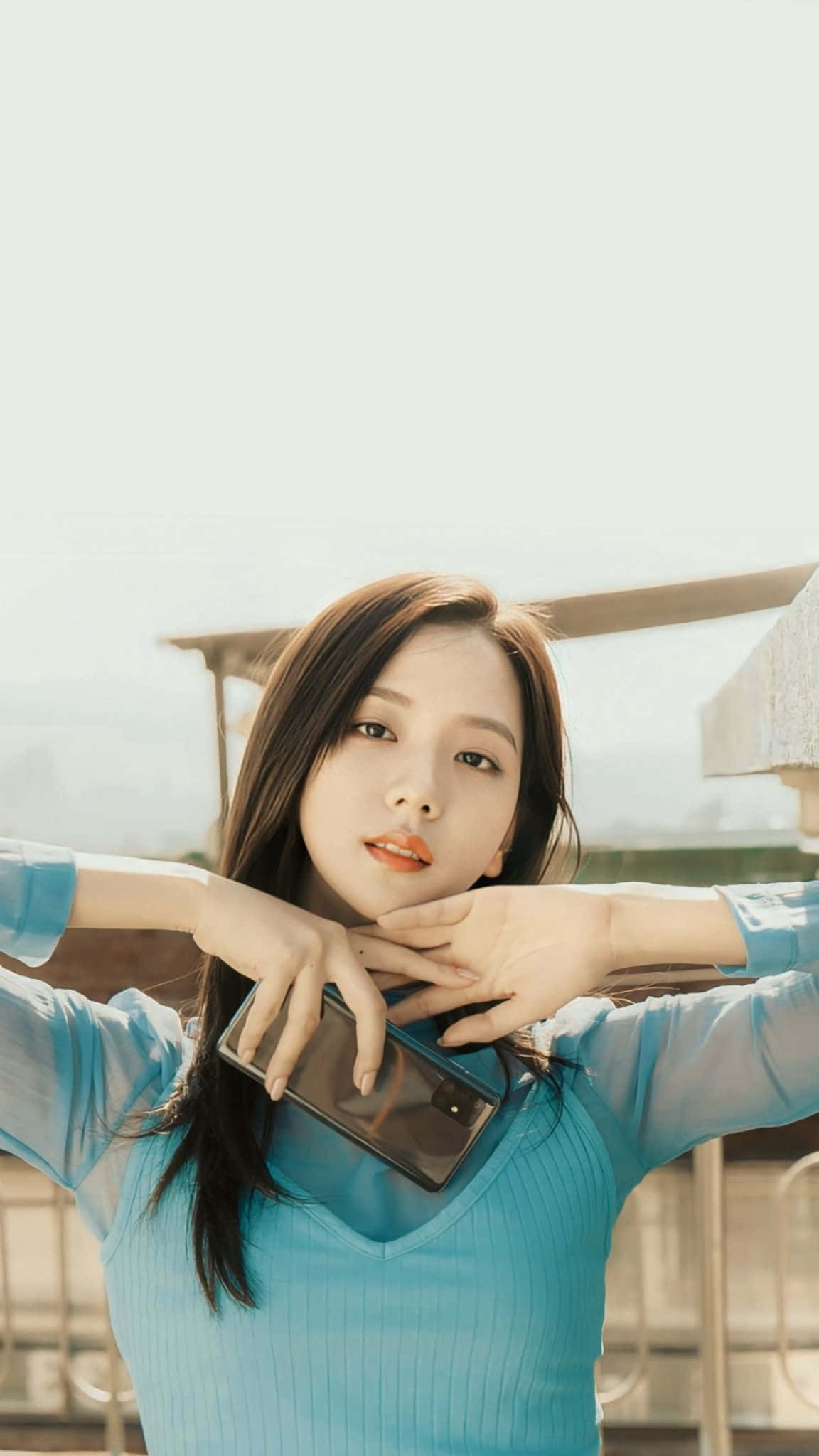 BLACKPINK 桌布 壁紙 — 一位身穿藍色羅紋上衣的年輕女性，在模糊的戶外背景下，手持電話，對著鏡頭露出溫柔的微笑。