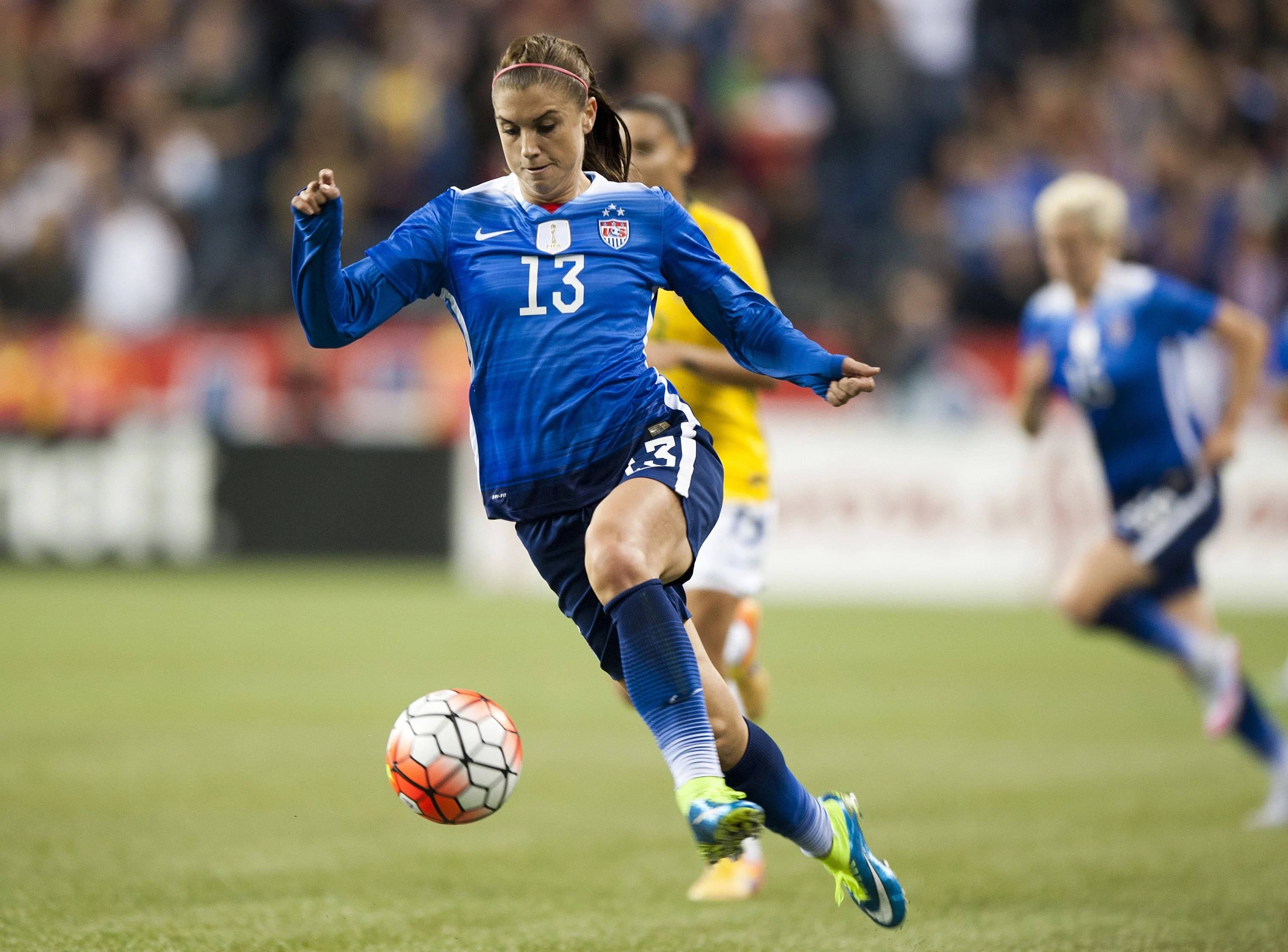 Alex Morgan 桌布 壁紙 — 一位身穿藍色美國13號球衣的女足球員在比賽中於綠色球場上踢一個色彩繽紛的球。