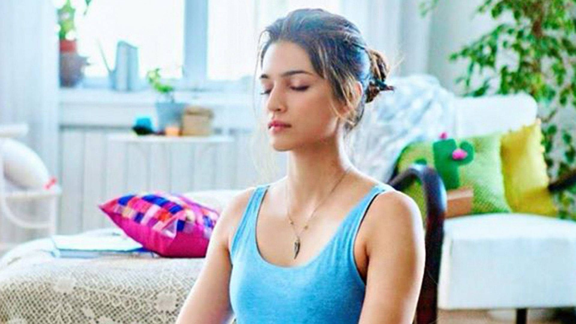 Kriti 桌布 壁紙 — 一位身穿藍色背心的女性，在陽光充足的房間裡，周圍有色彩繽紛的枕頭和綠色植物，平靜地坐著。
