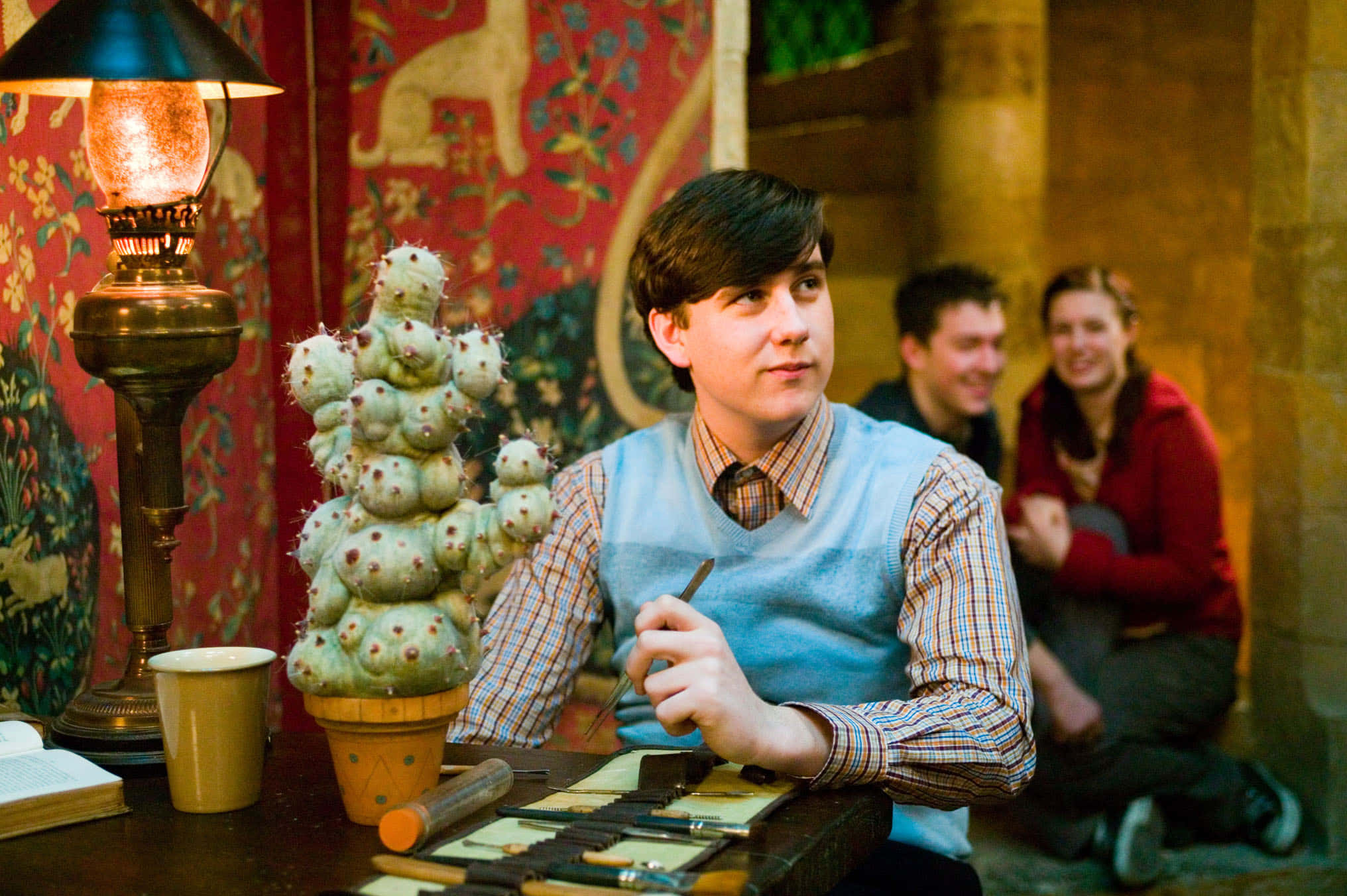 Neville Longbottom 桌布 壁紙 — 一位身穿藍色背心的年輕男子坐在書桌前，在溫暖的燈光下畫仙人掌，身後的朋友們在笑。