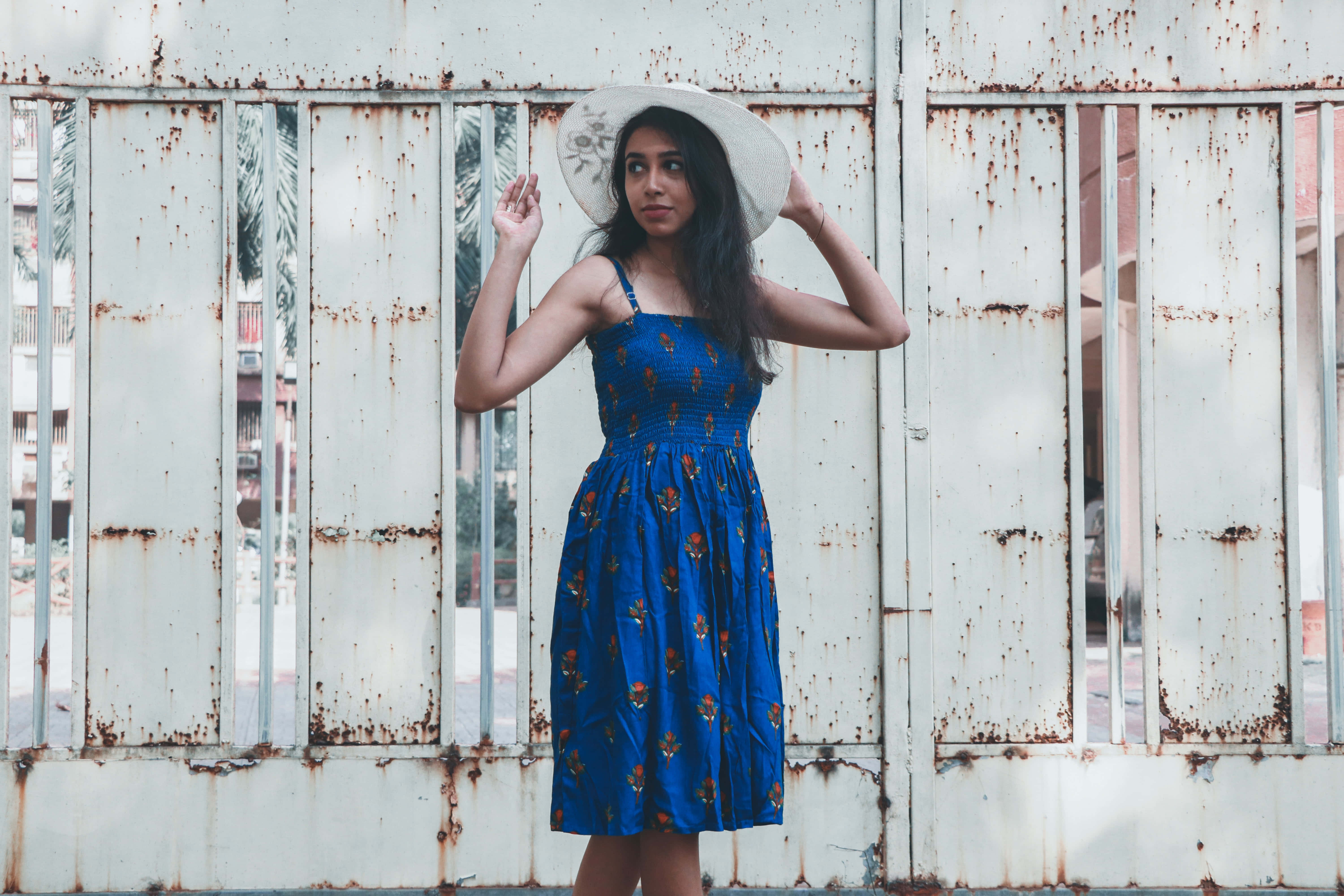 Navy Blue Dress 桌布 壁紙 — 一位身穿藍色花卉連衣裙和寬邊帽的女性，在風化的生鏽金屬柵欄前擺姿勢。