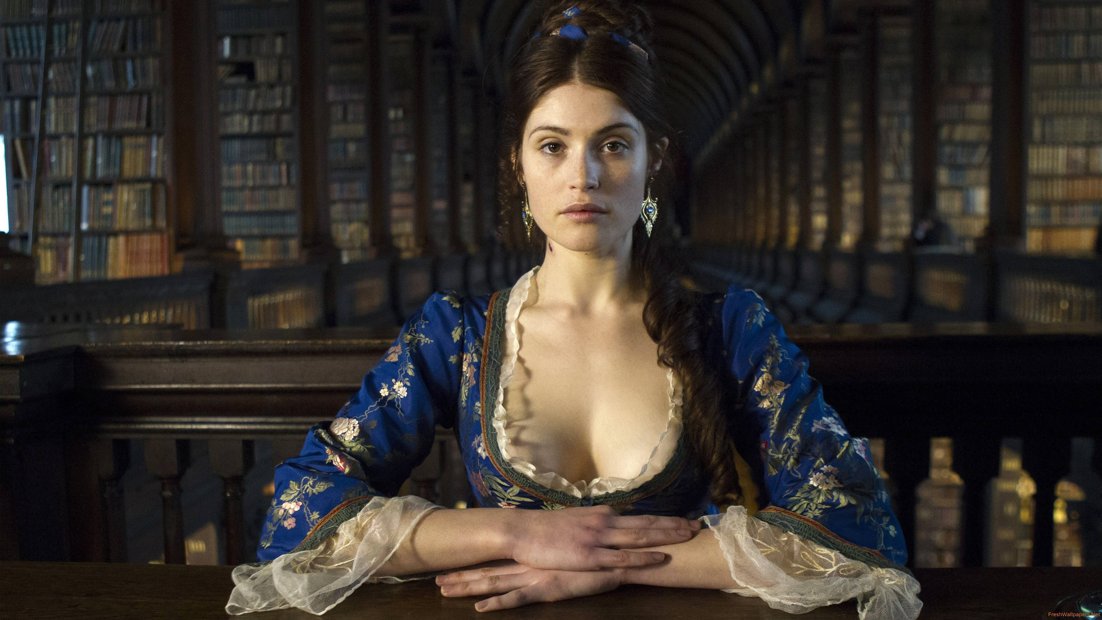 Gemma Arterton 桌布 壁紙 — 一位身穿藍色花卉長袍的女性坐在一個擺滿高聳書架的大圖書館的木桌旁。