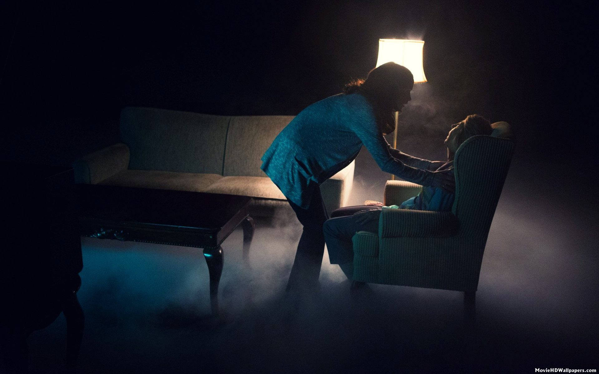 Insidious 桌布 壁紙 — 一位身穿藍色衣服的女性俯身靠近一個坐著的人，房間煙霧瀰漫、燈光昏暗，僅由一盞燈照亮，投射出戲劇性的陰影。