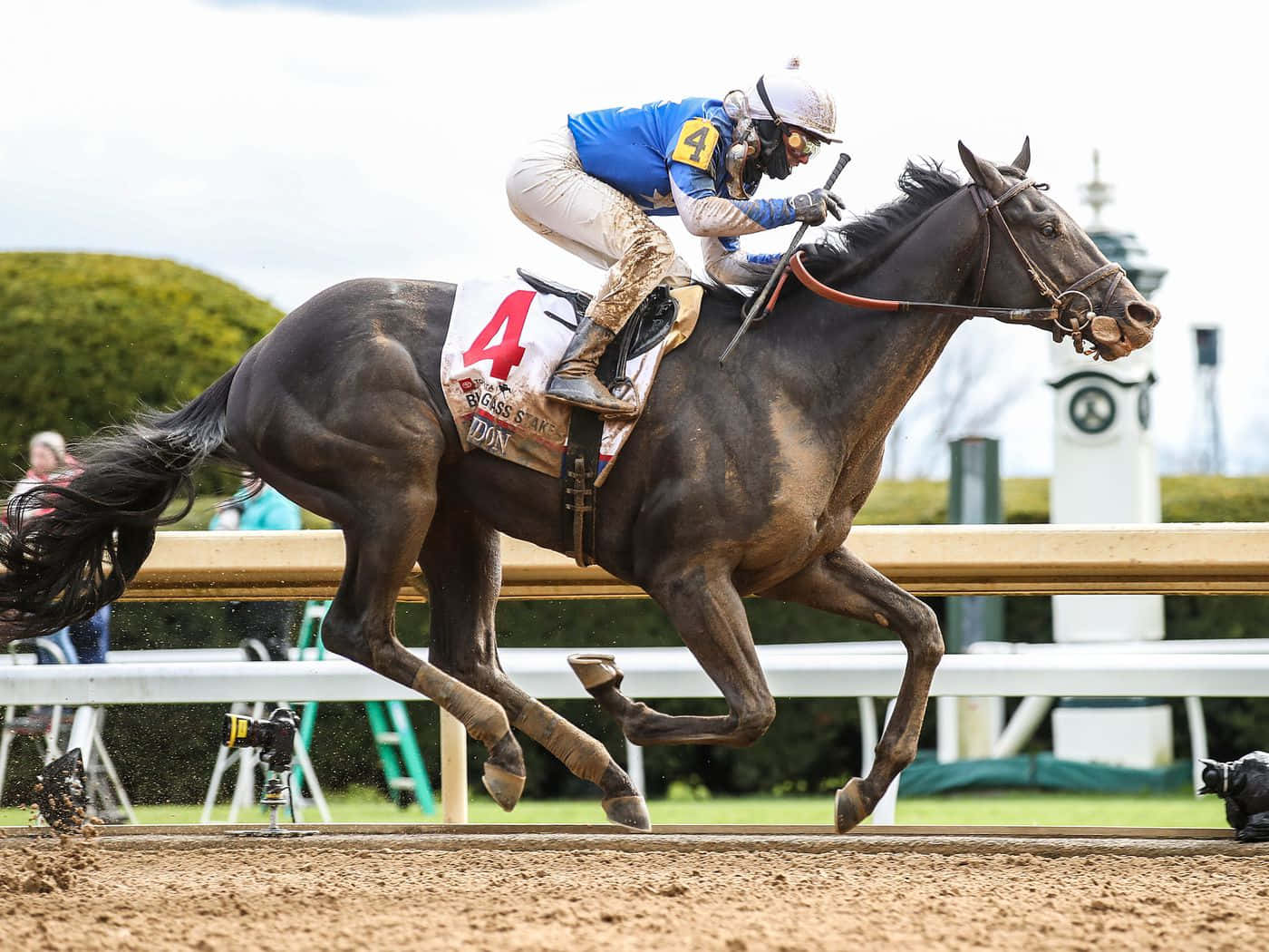 Kentucky Derby 桌布 壁紙 — 一位身穿藍色衣服的騎師向前傾，他的黑馬在泥地上強勁奔馳，背景有一座鐘樓。
