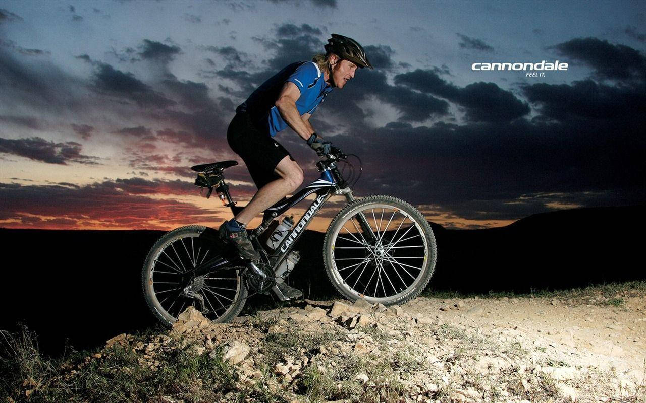 Tour 桌布 壁紙 — 一位身穿藍色裝備的自行車手在日落時分，騎著Cannondale登山車穿越岩石地形，天空中有戲劇性的雲彩。