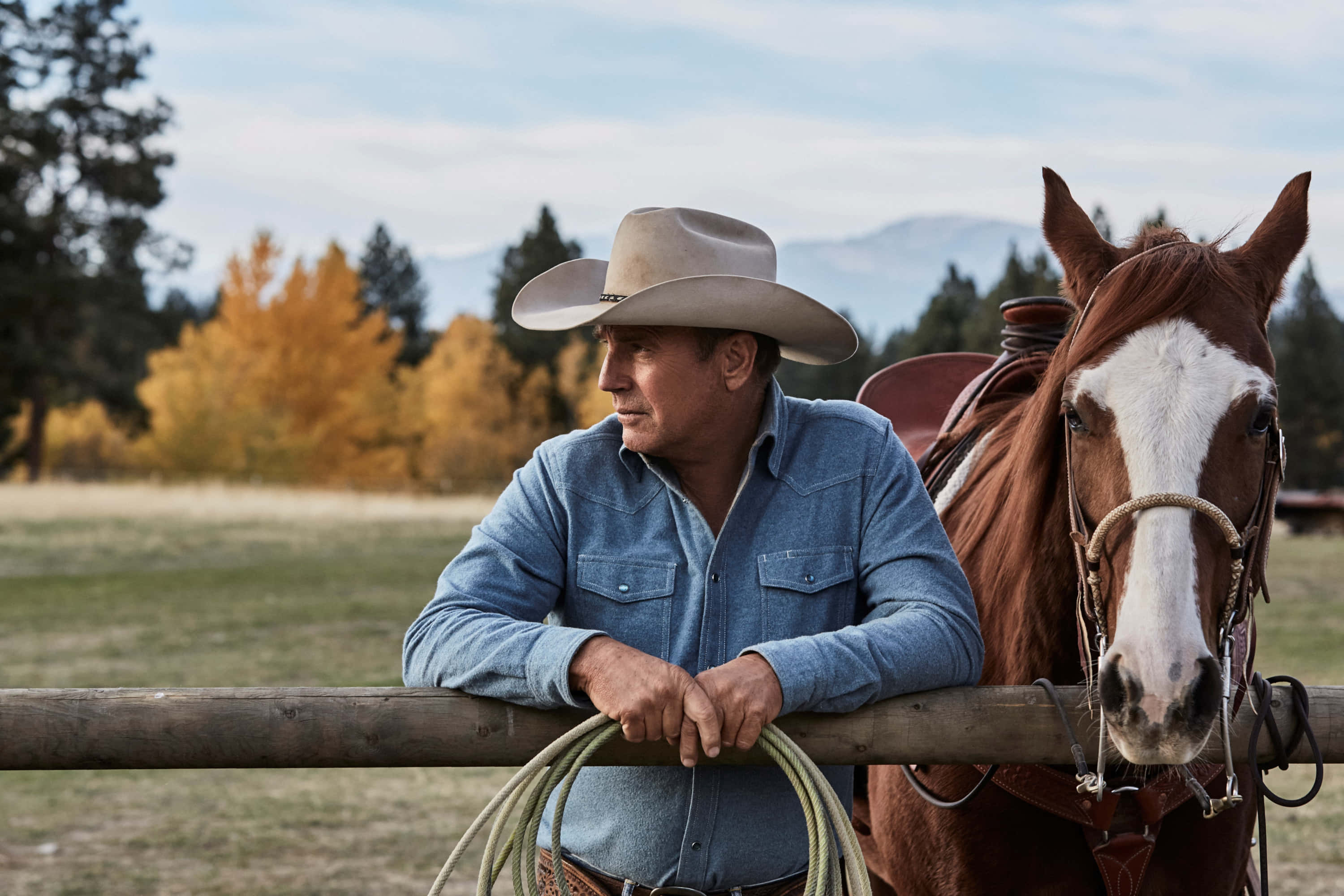 Yellowstone 桌布 壁紙 — 一位身穿藍色襯衫、戴帽子的牛仔靠著木柵欄，旁邊有一匹棕白相間的馬，背景是秋天的樹木和山脈。