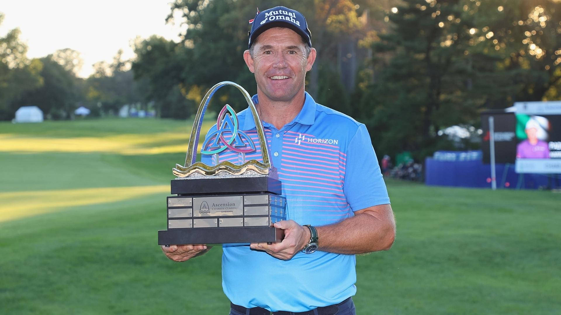 Trophy 桌布 壁紙 — 一位身穿藍色襯衫、面帶微笑的高爾夫球手，在陽光明媚的高爾夫球場上，背景有樹木，自豪地高舉一個華麗的大型獎盃。