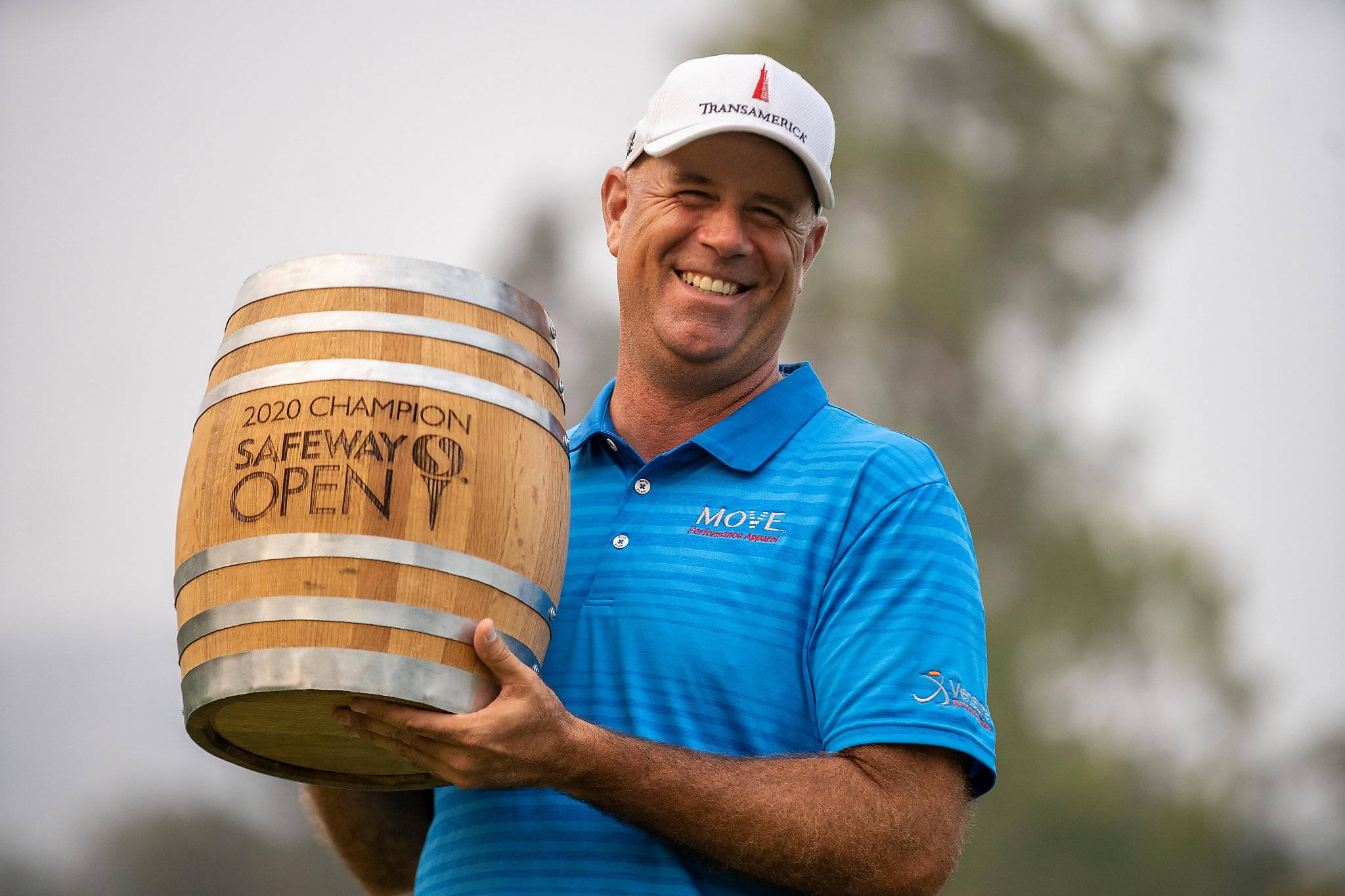Champion 桌布 壁紙 — 一位身穿藍色襯衫、面帶微笑的高爾夫球手，在模糊的戶外背景下，自豪地舉著一個刻有「2020 Champion Safeway Open」字樣的木桶獎盃。