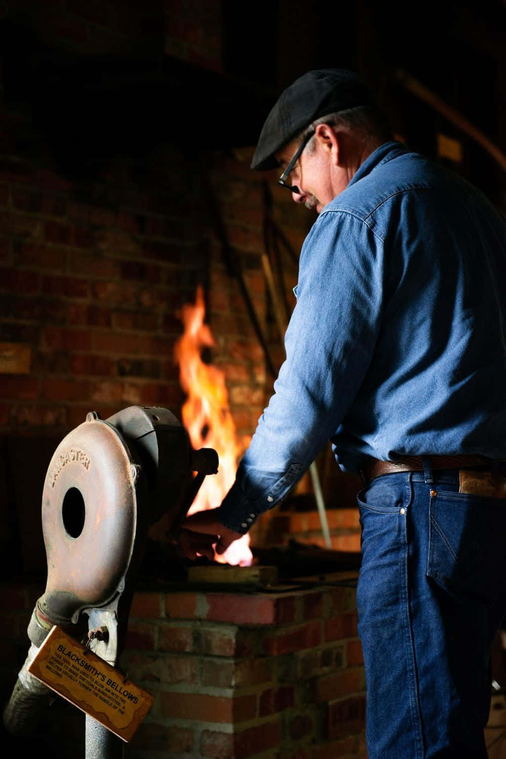 Blacksmith 桌布 壁紙 — 一位身穿藍色襯衫和帽子的鐵匠，在磚砌鍛造爐旁用風箱照料著熾熱的金屬。