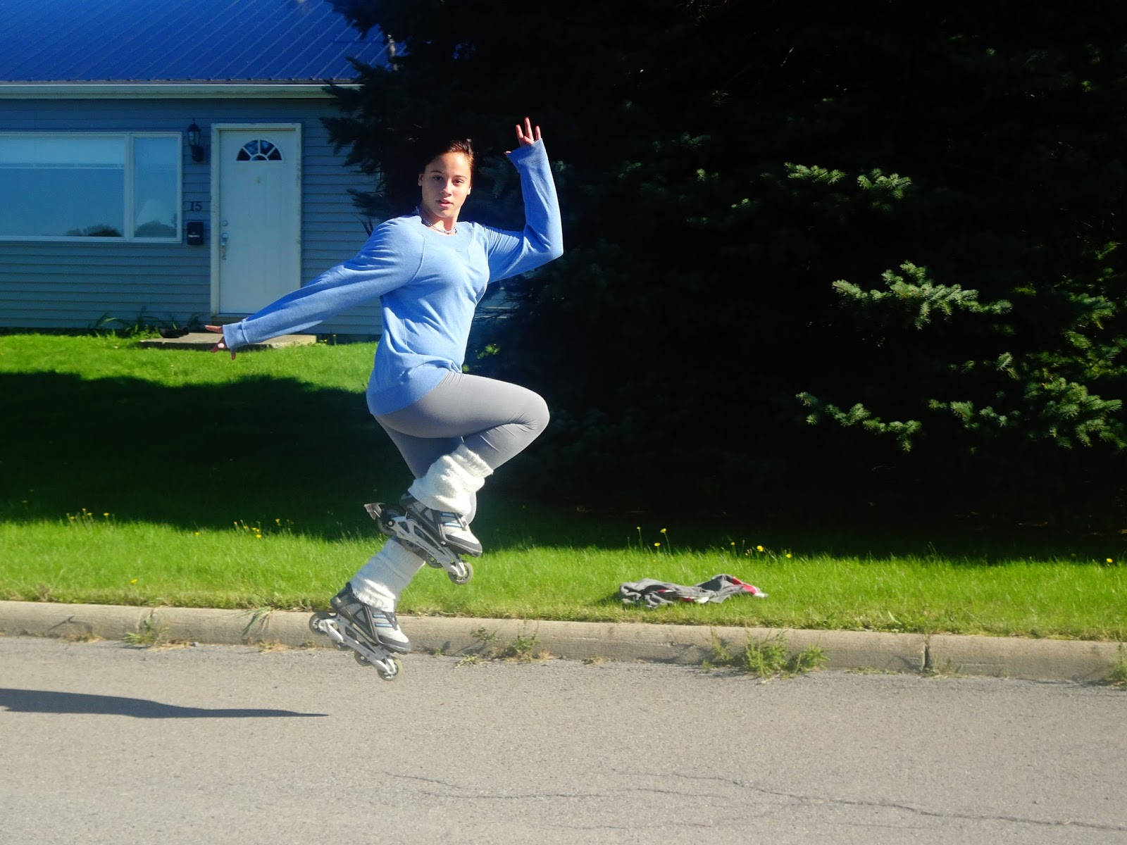skating 桌布 壁紙 — 一位身穿藍色襯衫和灰色褲子的女性，在藍色屋頂和綠色草坪的房子前，穿著直排輪鞋在空中跳躍。