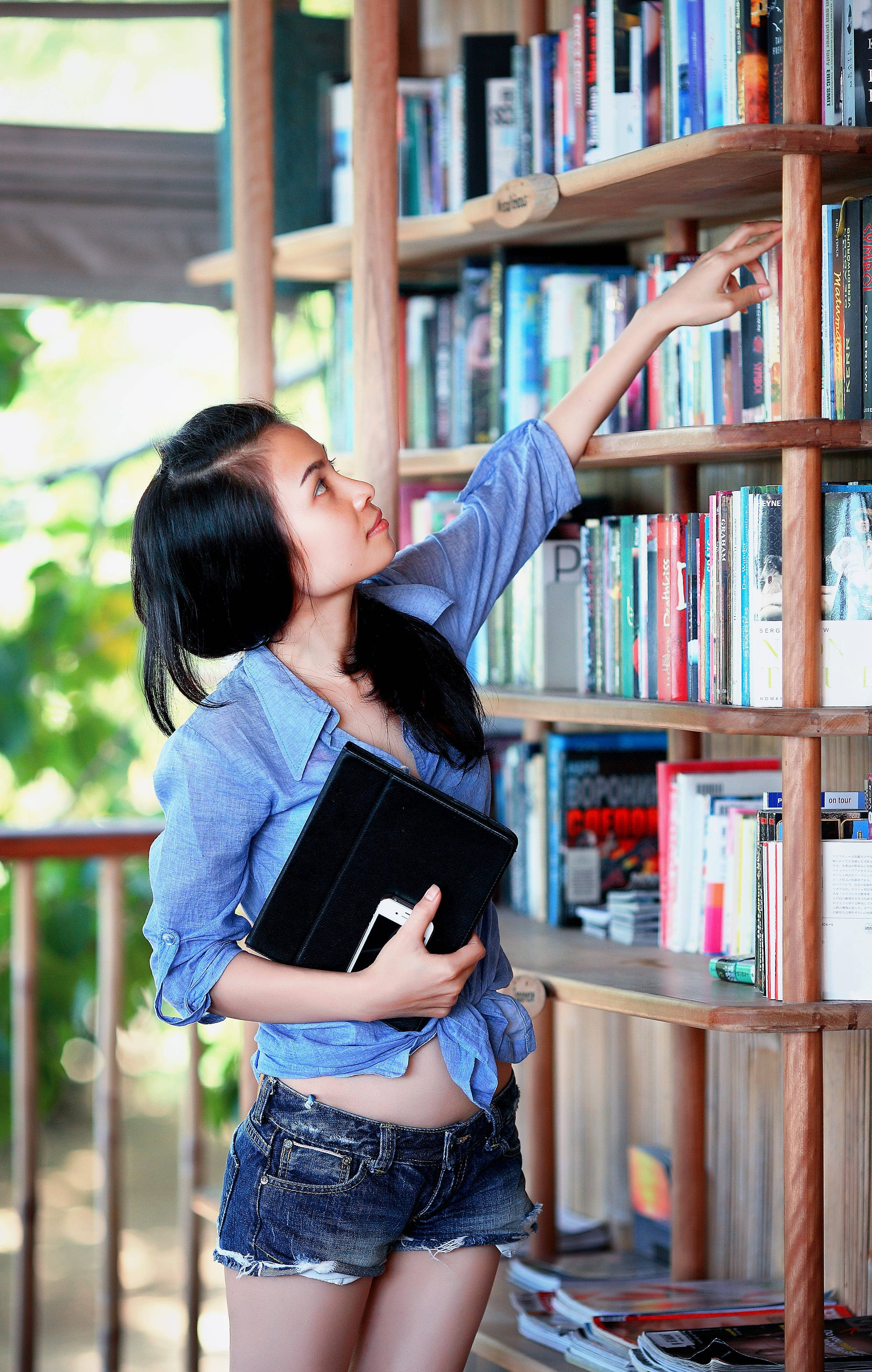 Reading 桌布 壁紙 — 一位身穿藍色襯衫和牛仔短褲的年輕女子，拿著一個黑色文件夾，伸手去拿木架上的書。