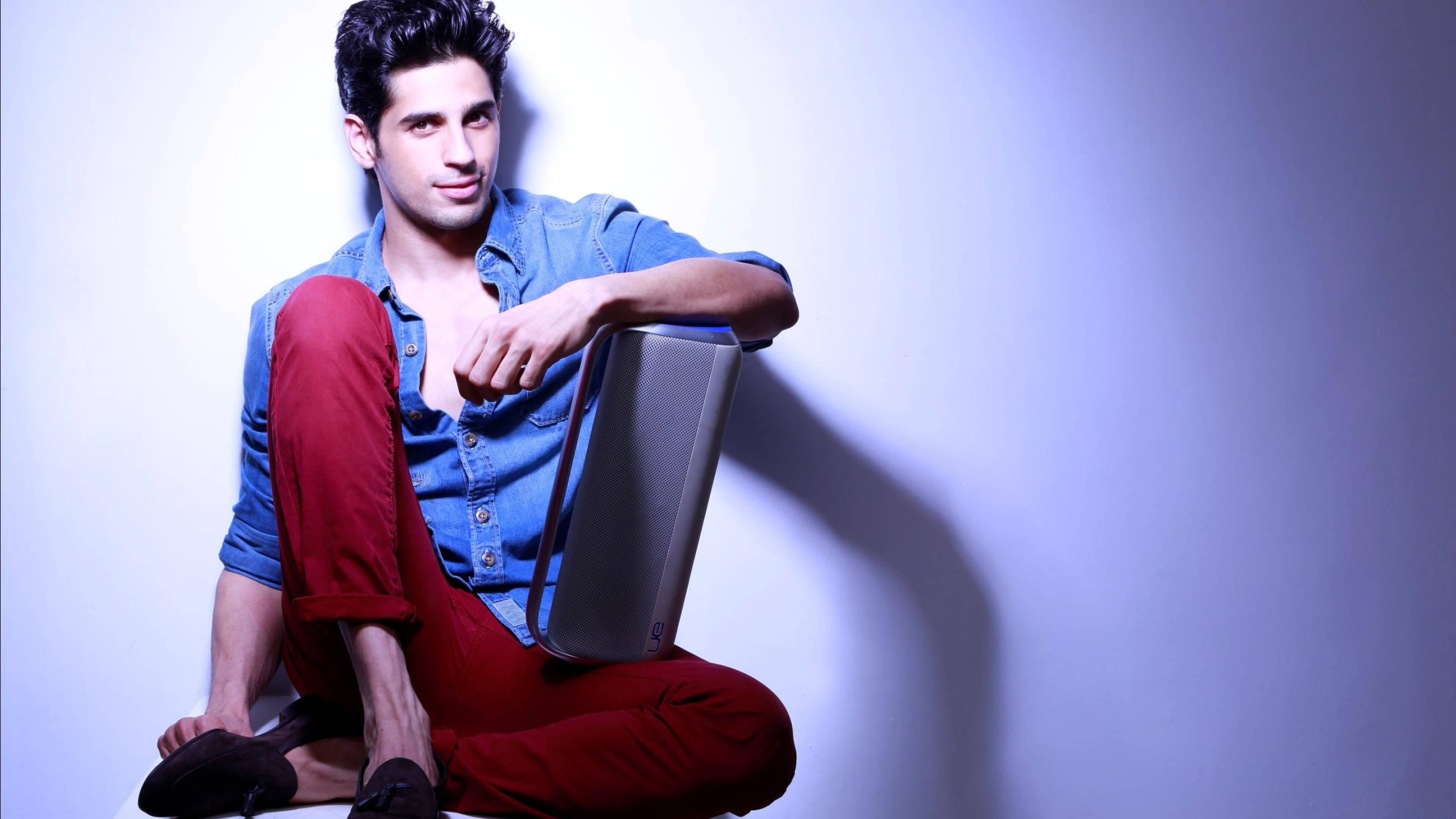 Sidharth Malhotra 桌布 壁紙 — 一位身穿藍色襯衫和紅色褲子的年輕男子，在柔和燈光的攝影棚背景下，隨意地坐著拿著筆記型電腦。