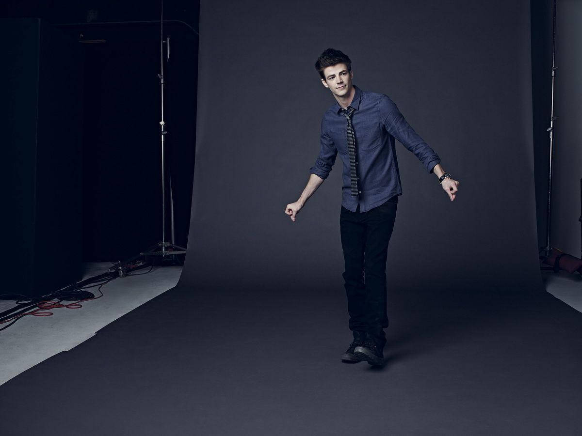 Grant Gustin 桌布 壁紙 — 一位身穿藍色襯衫和領帶的年輕男子自信地走過一個光線昏暗、背景深灰色且照明設備可見的攝影棚。