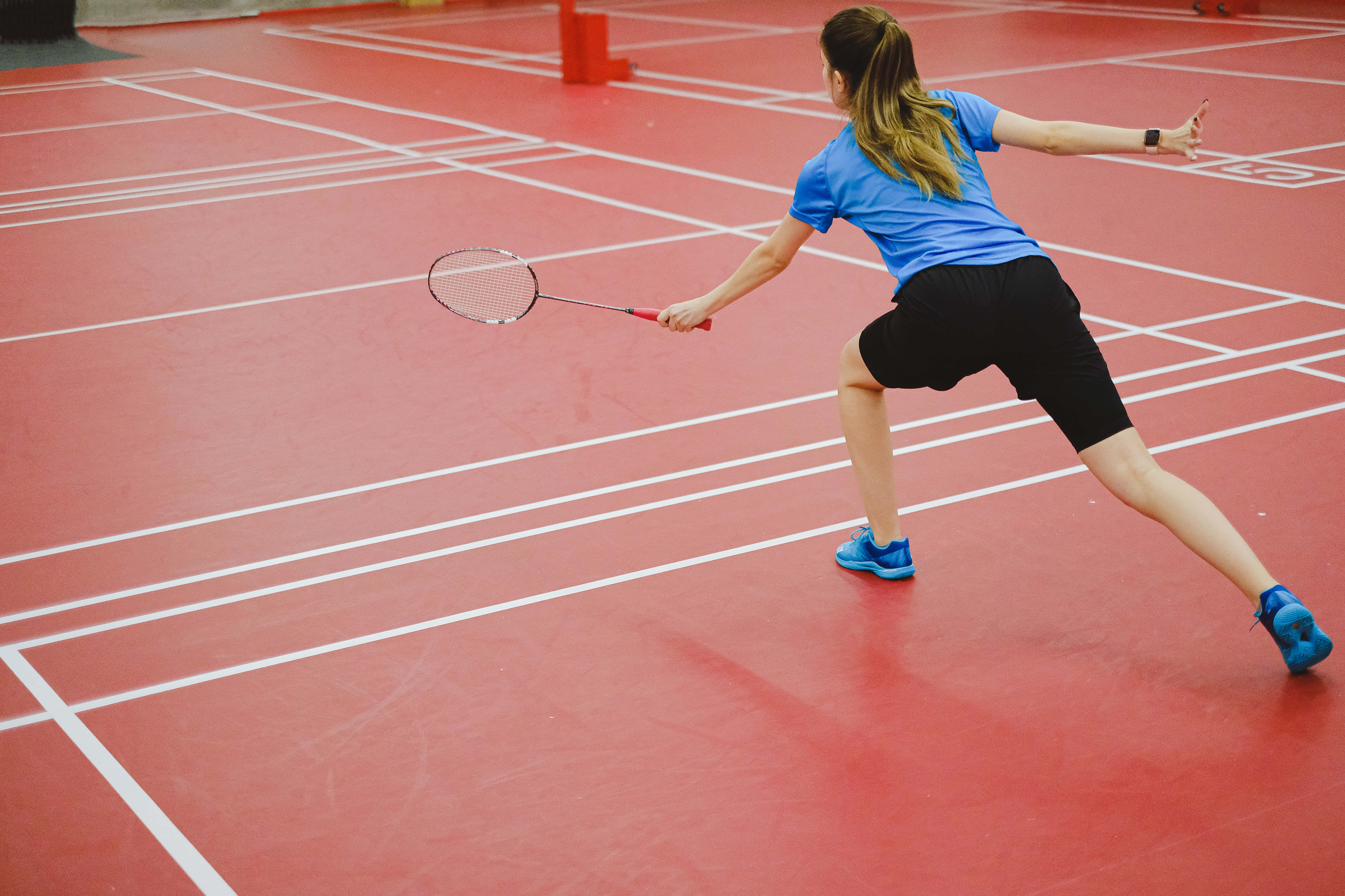 Badminton 桌布 壁紙 — 一位身穿藍色襯衫和黑色短褲的女羽球運動員在帶有白色線條的紅色場地上向前衝刺，揮動她的球拍。
