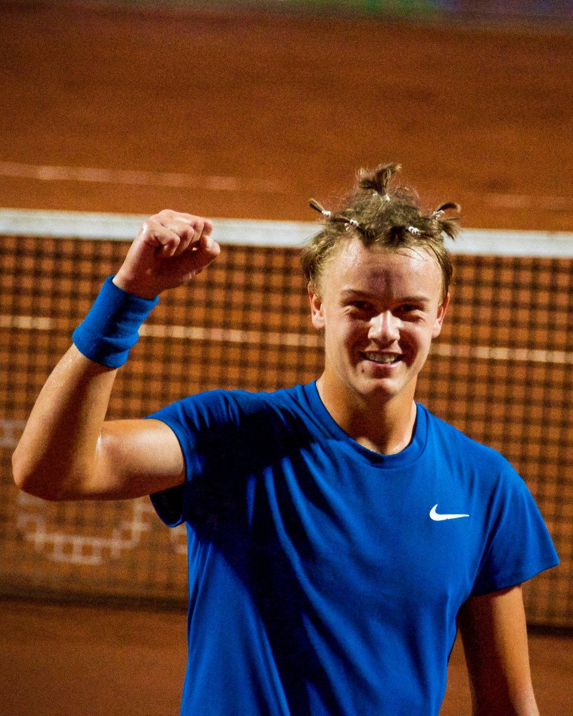 Tennis 桌布 壁紙 — 一位身穿藍色襯衫的勝利網球選手在體育場燈光下的紅土場上舉起拳頭慶祝勝利。