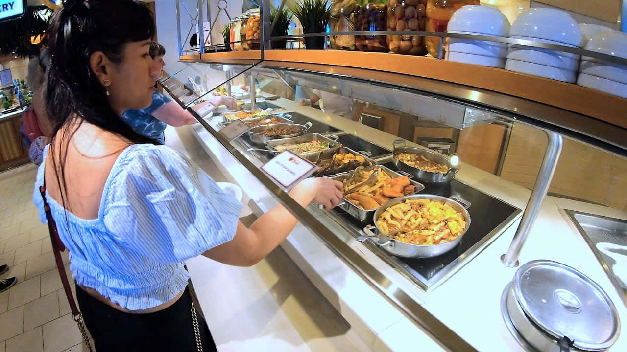 Carnival Conquest 桌布 壁紙 — 一位身穿藍色襯衫的女性正在明亮的自助餐檯前選擇食物，餐檯上擺滿了玻璃罩下熱氣騰騰的菜餚。