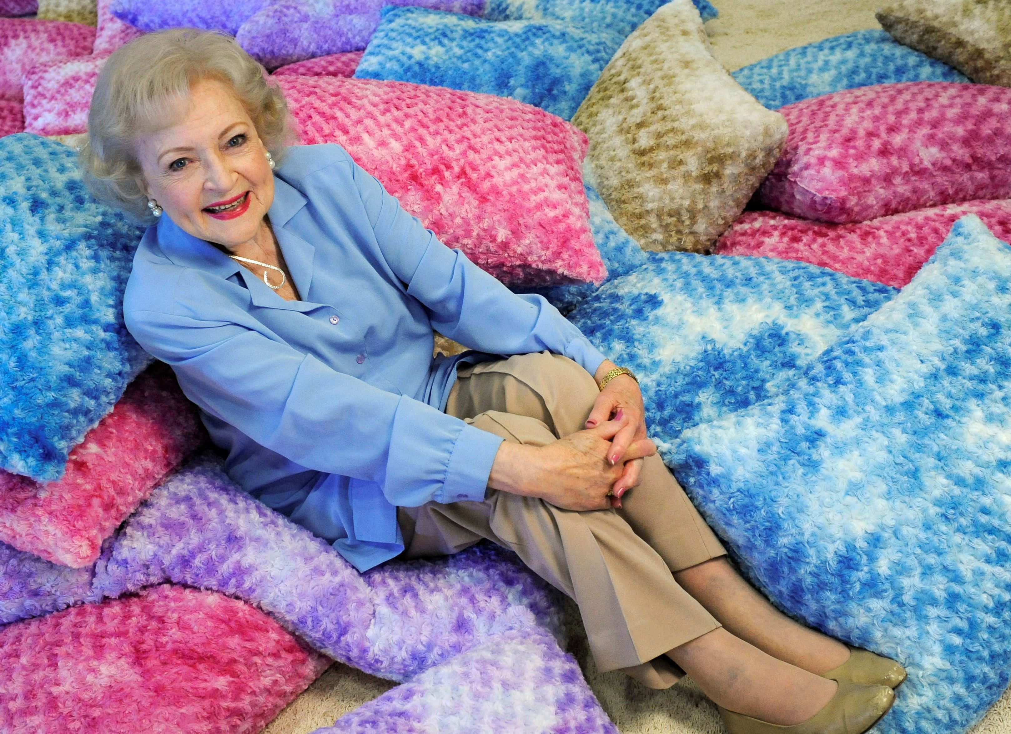 Betty White 桌布 壁紙 — 一位身穿藍色襯衫的年長女性在舒適的室內環境中，坐在色彩繽紛的柔軟枕頭之間微笑。
