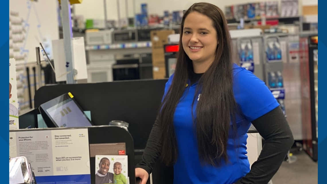 Best Buy 桌布 壁紙 — 一位身穿藍色襯衫的微笑女士站在光線充足的零售店的收銀台後面，背景是擺滿產品的貨架。