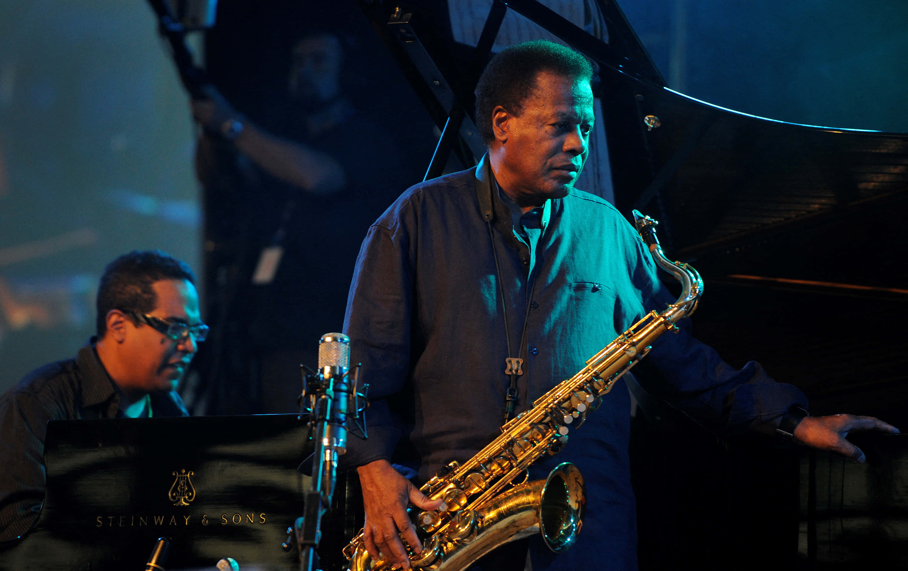 Wayne Shorter 桌布 壁紙 — 一位身穿藍色襯衫的爵士薩克斯風手在舞台燈光下演奏金色薩克斯風，背景有鋼琴和另一位音樂家。