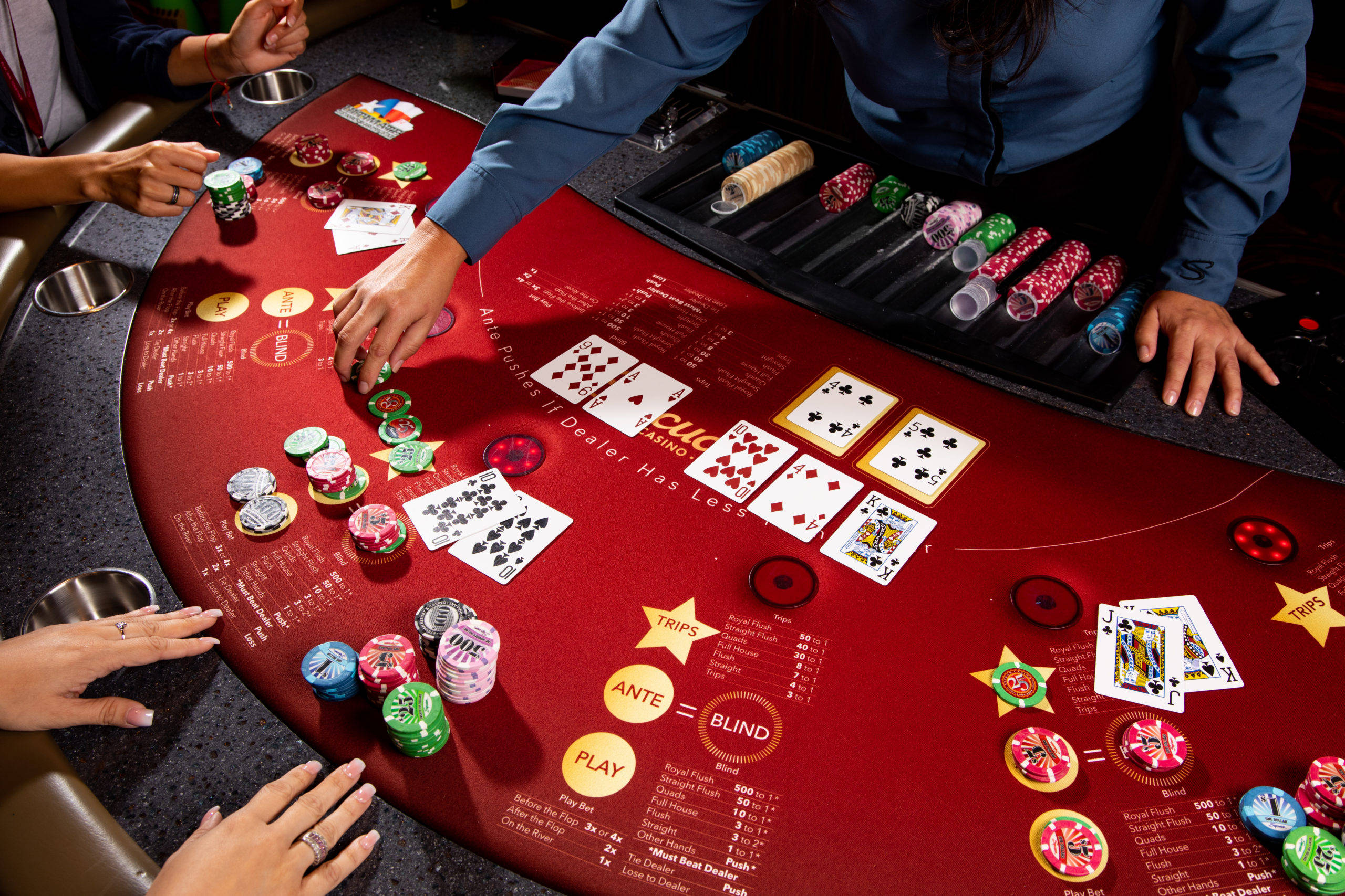 Poker 桌布 壁紙 — 一位身穿藍色襯衫的荷官管理著一張紅色撲克桌，玩家在激烈遊戲中放置彩色籌碼和紙牌。