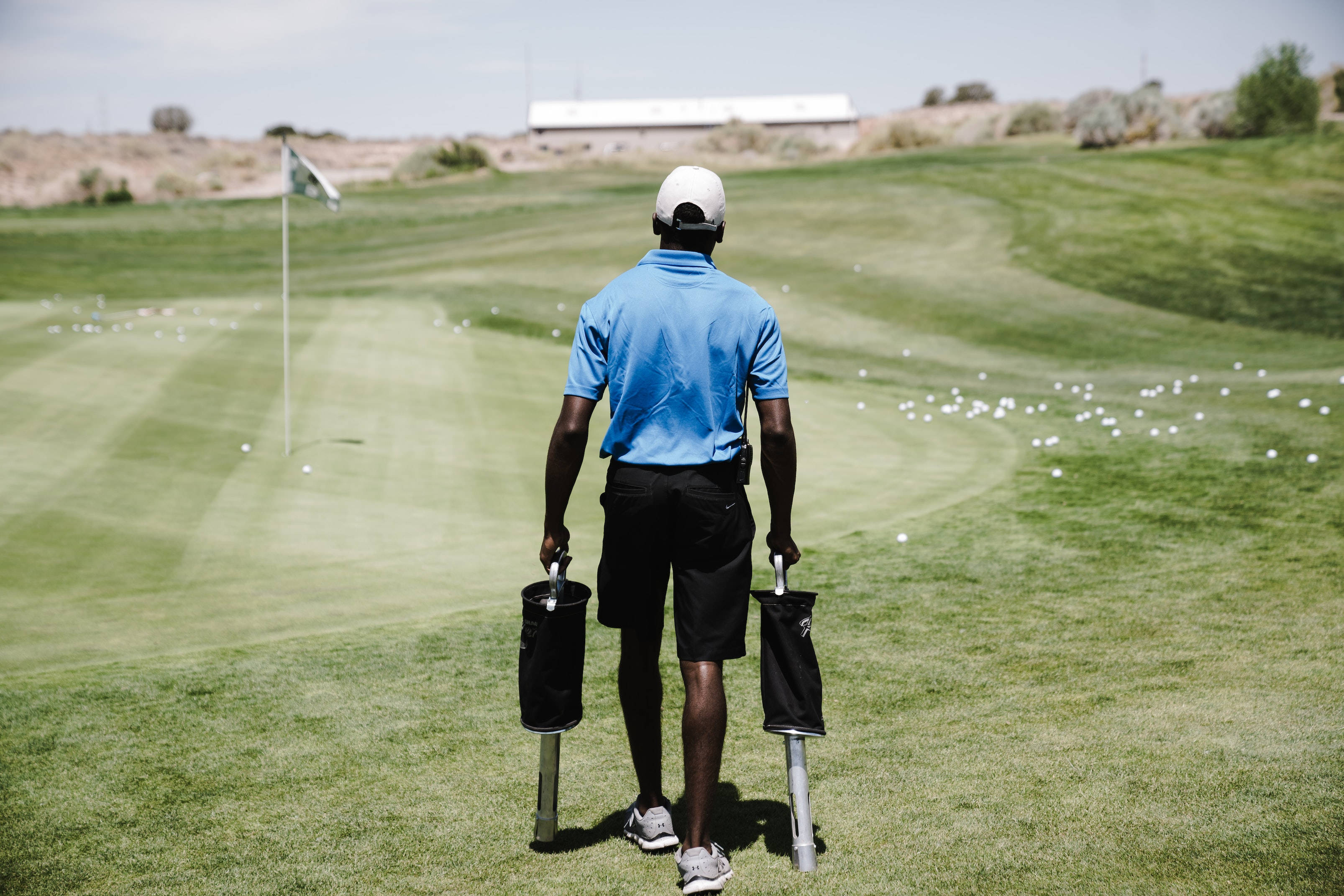 4K Golf 桌布 壁紙 — 一位身穿藍色襯衫的高爾夫球手在晴朗的天空下，帶著兩支球桿和一袋高爾夫球走向果嶺。
