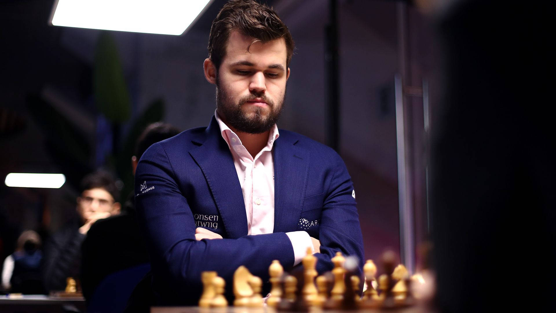 Magnus 桌布 壁紙 — 一位身穿藍色西裝、專注的棋手在比賽中思考下一步，背景有模糊的觀眾。