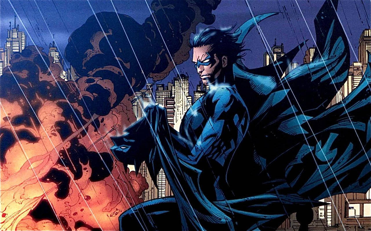 Nightwing 桌布 壁紙 — 一位身穿藍色西裝、披著飄逸斗篷的超級英雄，在雨水浸濕的城市景觀中挑釁地站立，身後爆發著熾熱的爆炸。