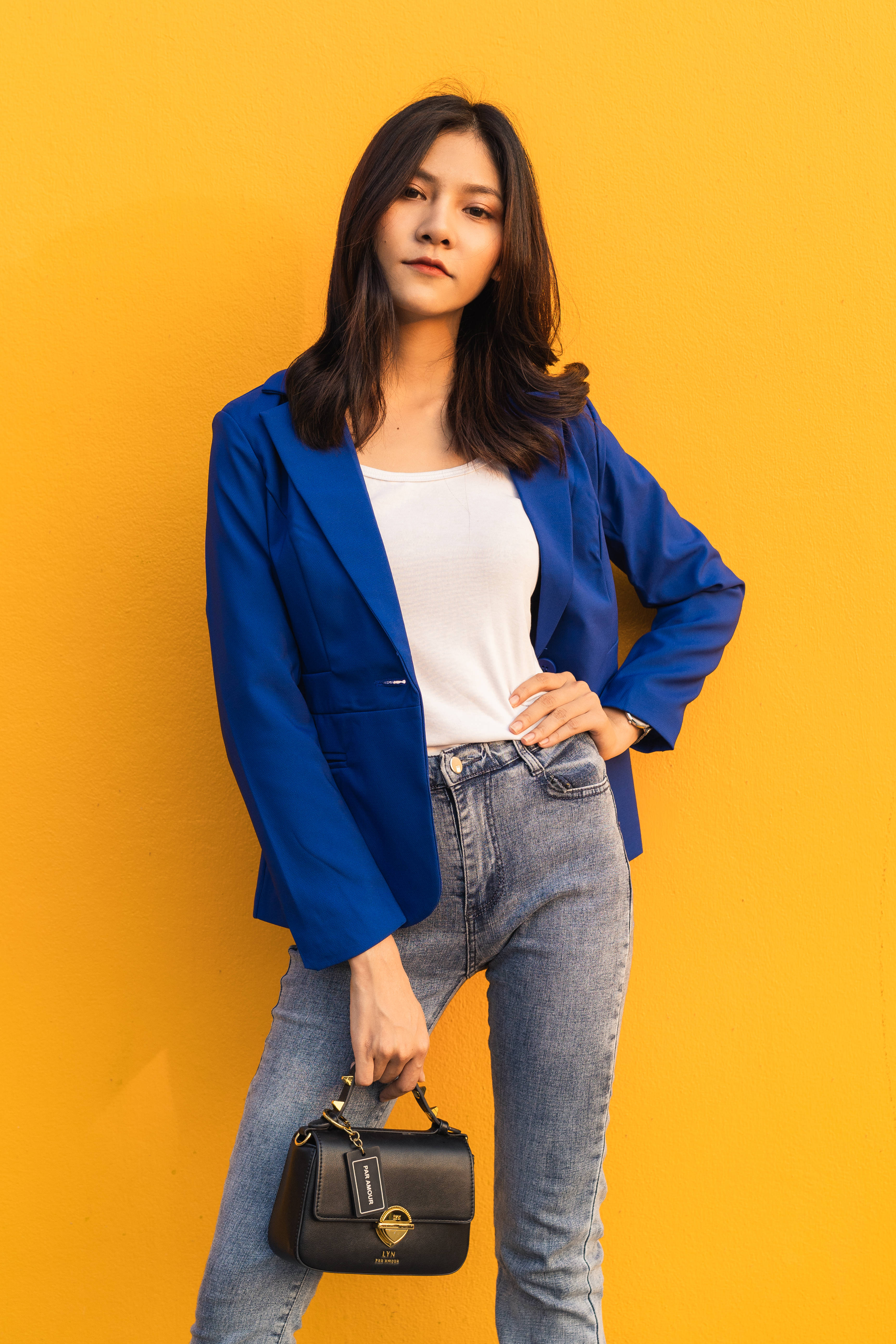 酷女孩 桌布 壁紙 — 一位身穿藍色西裝外套和牛仔褲的女性，自信地站在鮮黃色的牆壁前，手持黑色手提包。