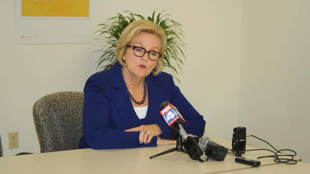 政治 桌布 壁紙 — 一位身穿藍色西裝外套的女性在記者會桌上對著FOX 4麥克風講話，身後有一盆盆栽。