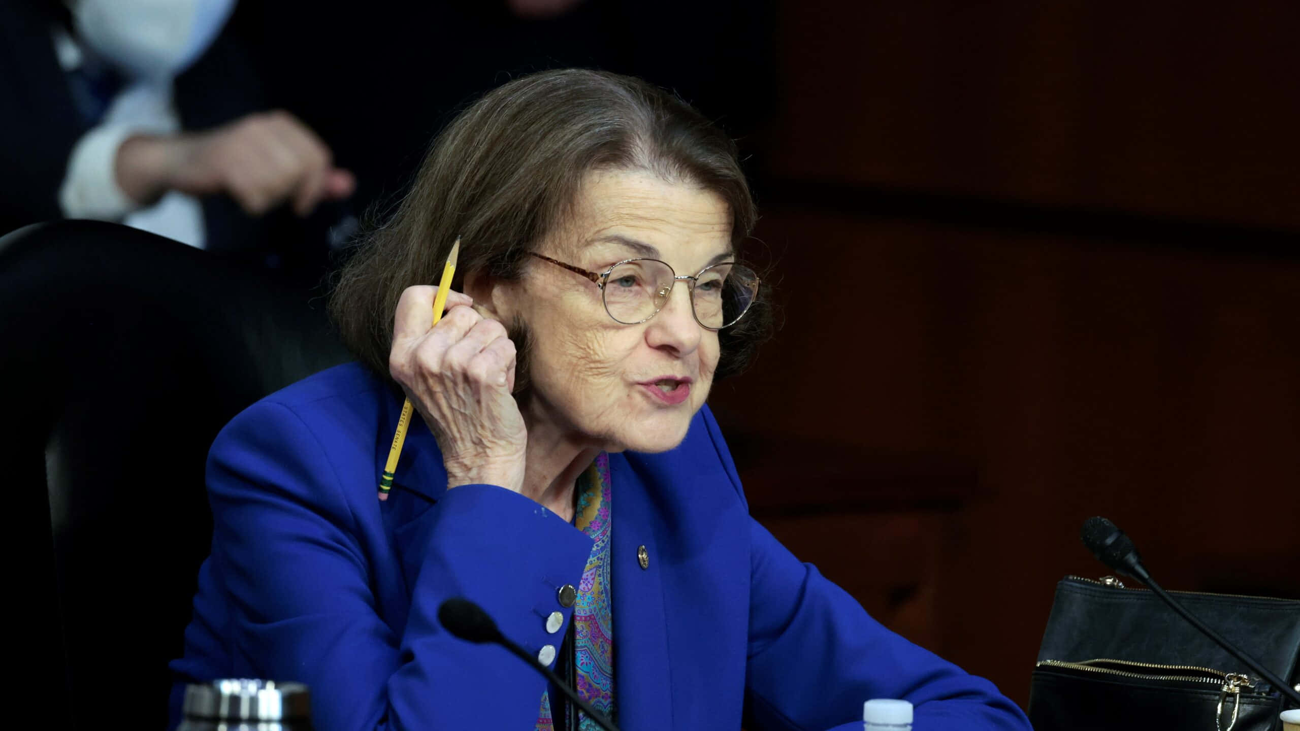 Dianne Feinstein 桌布 壁紙 — 一位身穿藍色西裝外套的老年女性在正式聽證會上發言時，將一支黃色鉛筆放在耳邊。
