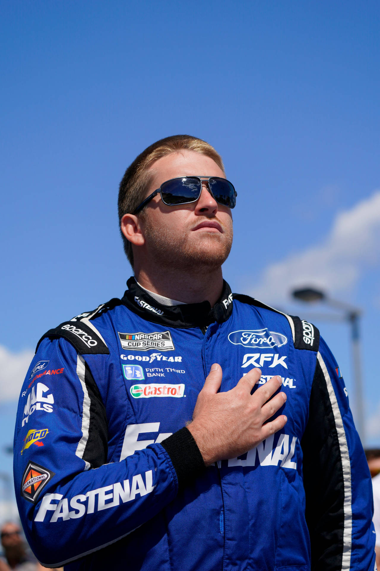  壁紙 — 一位身穿藍色賽車服的NASCAR車手在晴朗的藍天下莊嚴地將手放在心口上。