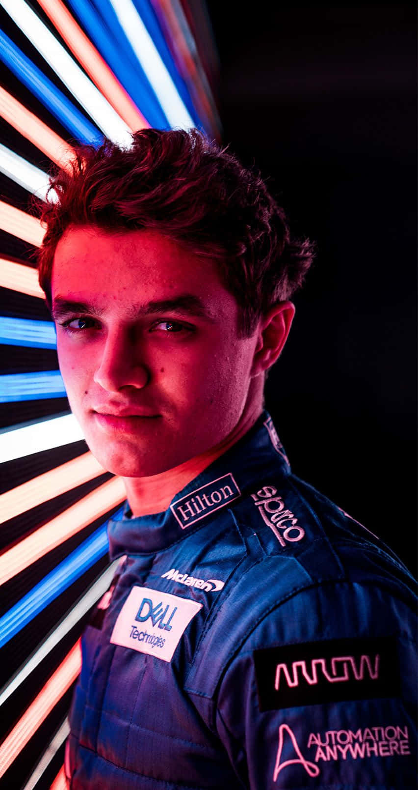 lando norris 桌布 壁紙 — 一位身穿藍色賽車服的年輕賽車手站在充滿活力的霓虹燈背景前，背景有紅色和藍色的條紋。