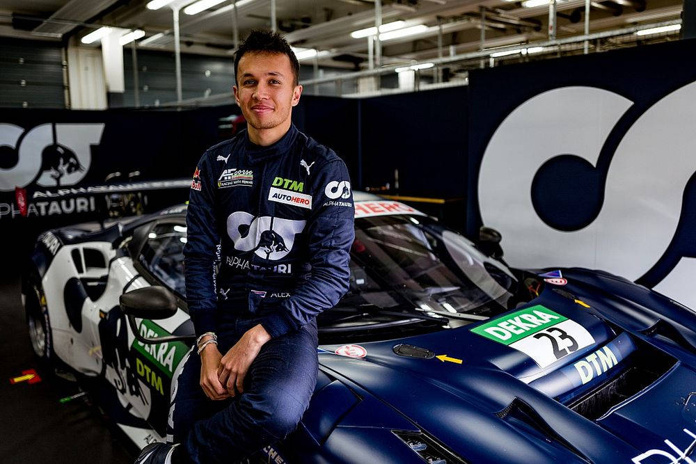 Racing 桌布 壁紙 — 一位身穿藍色車隊外套的賽車手自信地坐在車庫內一輛時尚藍白相間的DTM賽車引擎蓋上。
