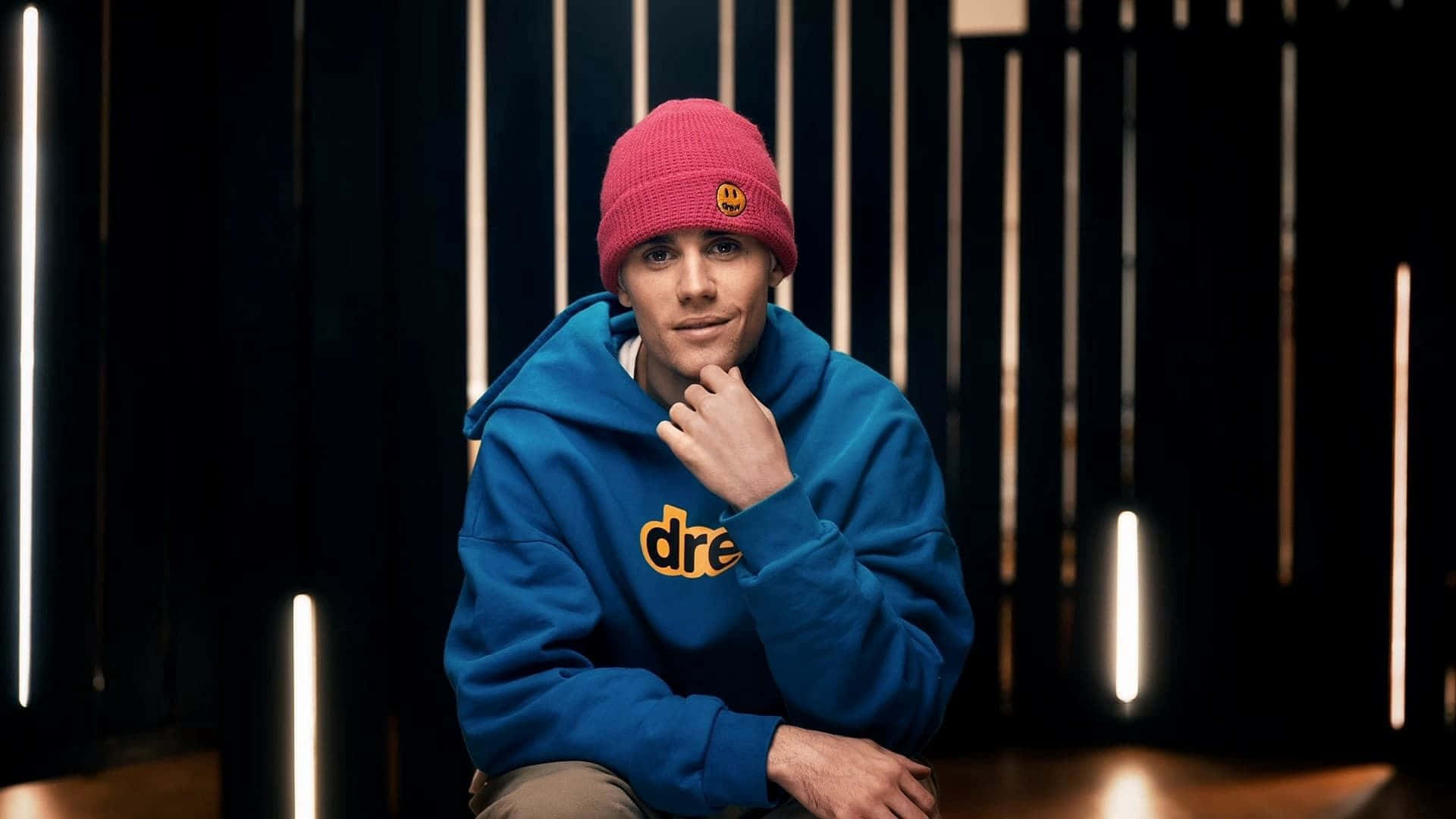 Justin Bieber 2021 桌布 壁紙 — 一位身穿藍色連帽衫和紅色毛帽的年輕男子，在帶有垂直發光燈光的深色背景前若有所思地擺姿勢。