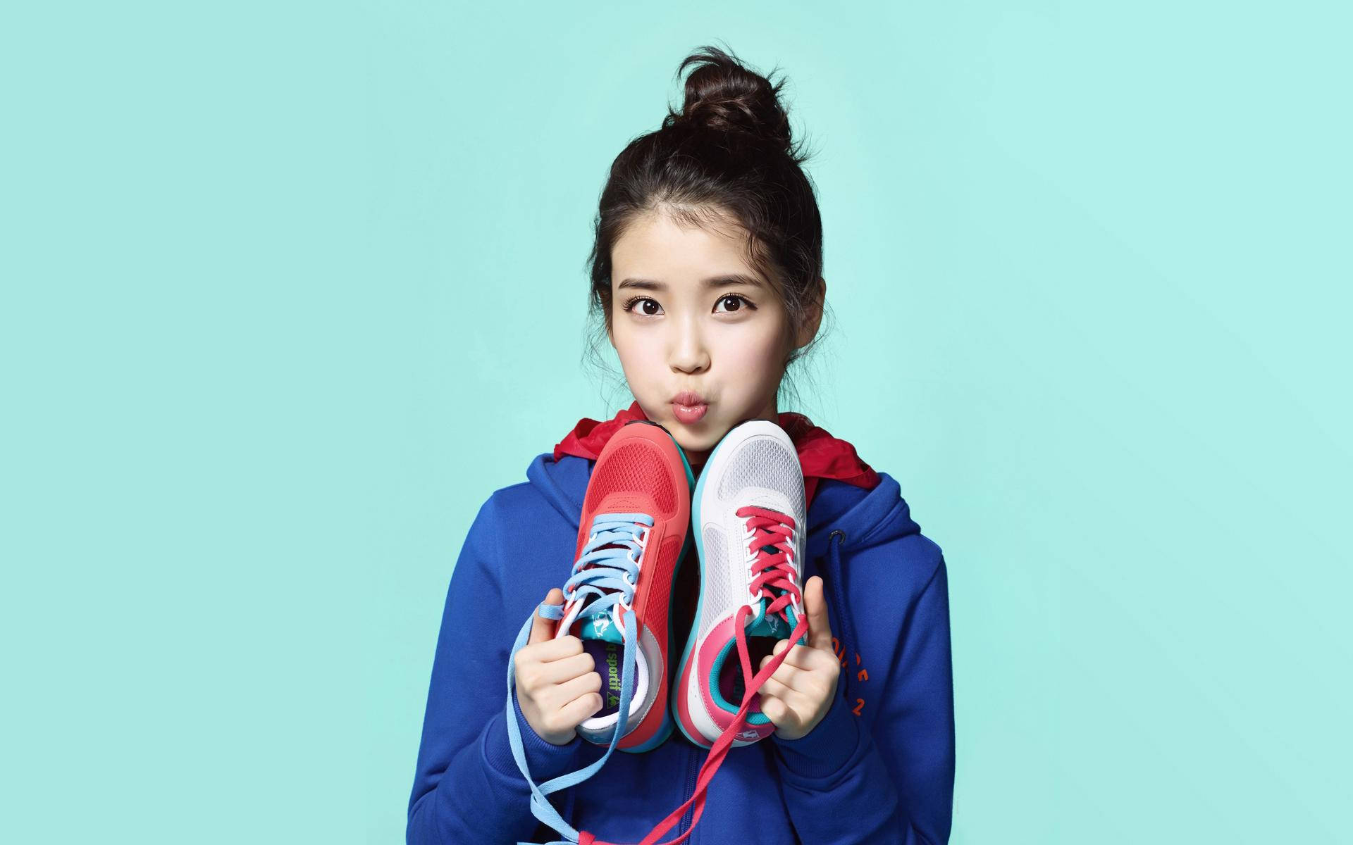 Kpop Aesthetic 桌布 壁紙 — 一位身穿藍色連帽衫的年輕女子，在純青色背景前舉起兩雙色彩繽紛的運動鞋。