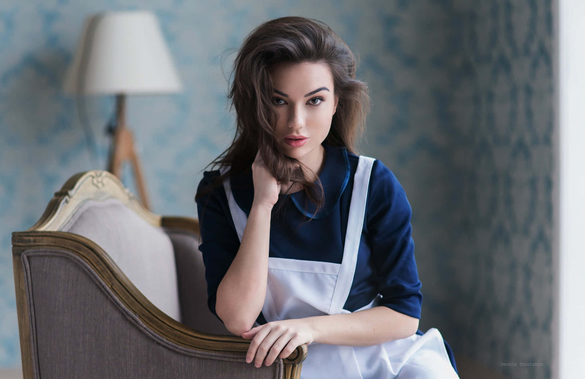Navy Blue Shirt 桌布 壁紙 — 一位身穿藍色連衣裙和白色圍裙的女性，坐在裝飾華麗的椅子上，背後是帶圖案的藍色壁紙，若有所思。