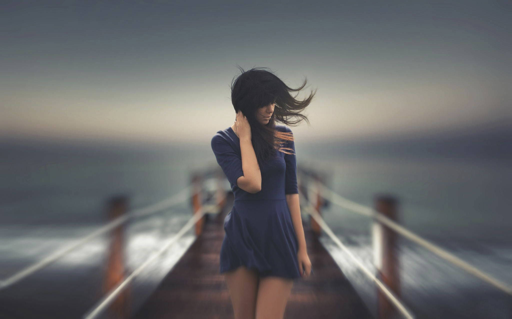 windy 桌布 壁紙 — 一位身穿藍色連衣裙的女性站在木製碼頭上，風吹動她的頭髮，背景是柔和的黃昏天空。