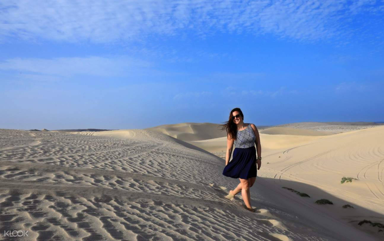 Qatar 桌布 壁紙 — 一位身穿藍色連衣裙的女性赤腳走在紋理沙丘上，頭頂是廣闊的藍天，飄著縷縷薄雲。
