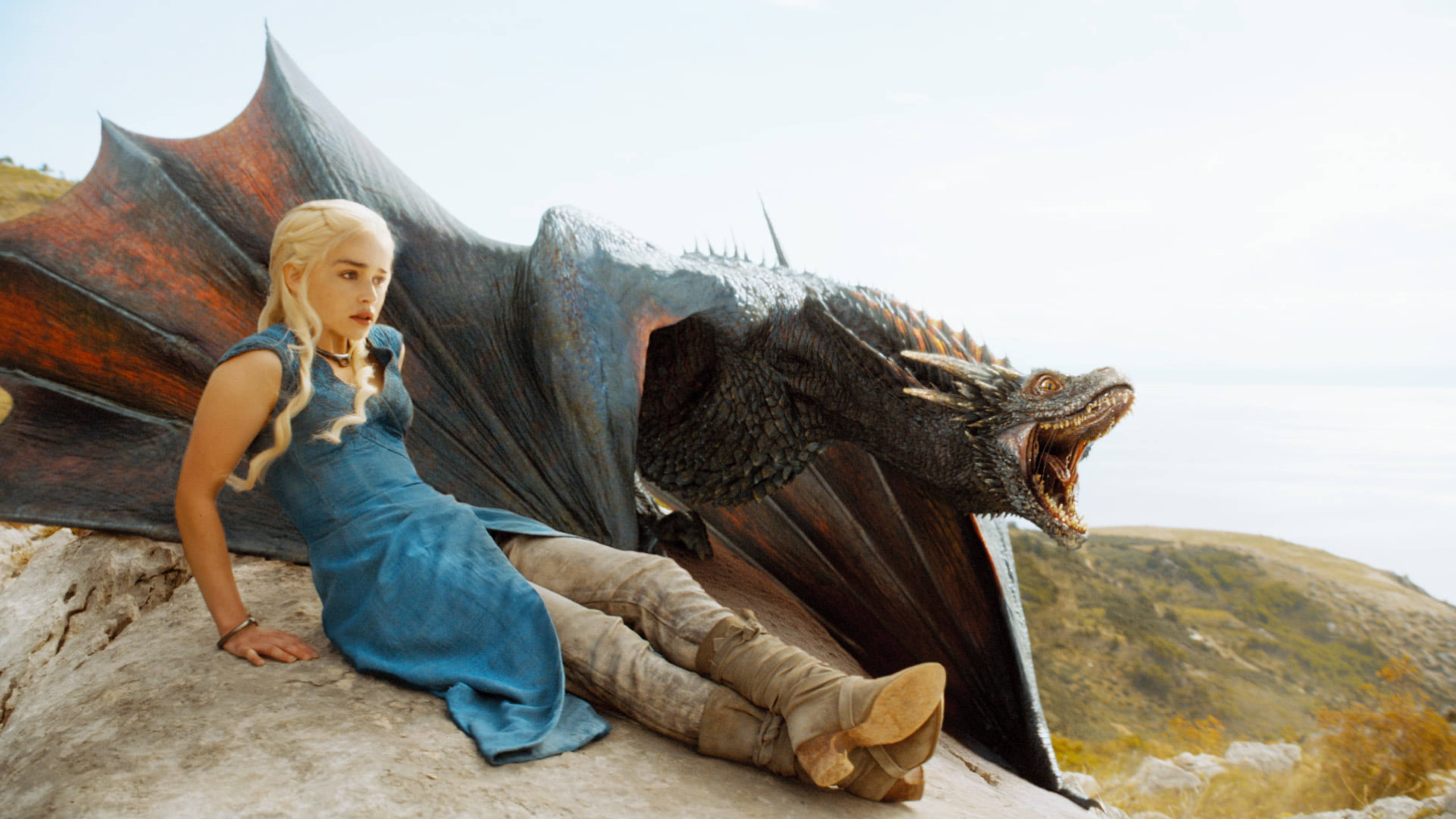 daenerys 桌布 壁紙 — 一位身穿藍色連衣裙的女性靠在一隻展開翅膀的巨大黑色龍旁邊，位於俯瞰大海的岩石懸崖上。