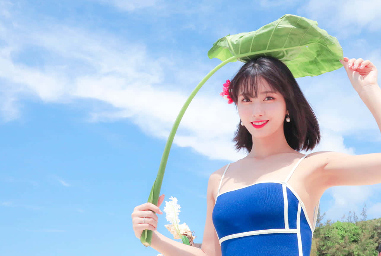 twice 桌布 壁紙 — 一位身穿藍色連衣裙的微笑女子，在明亮的藍天和蓬鬆雲朵下，將一片巨大的綠葉舉過頭頂。