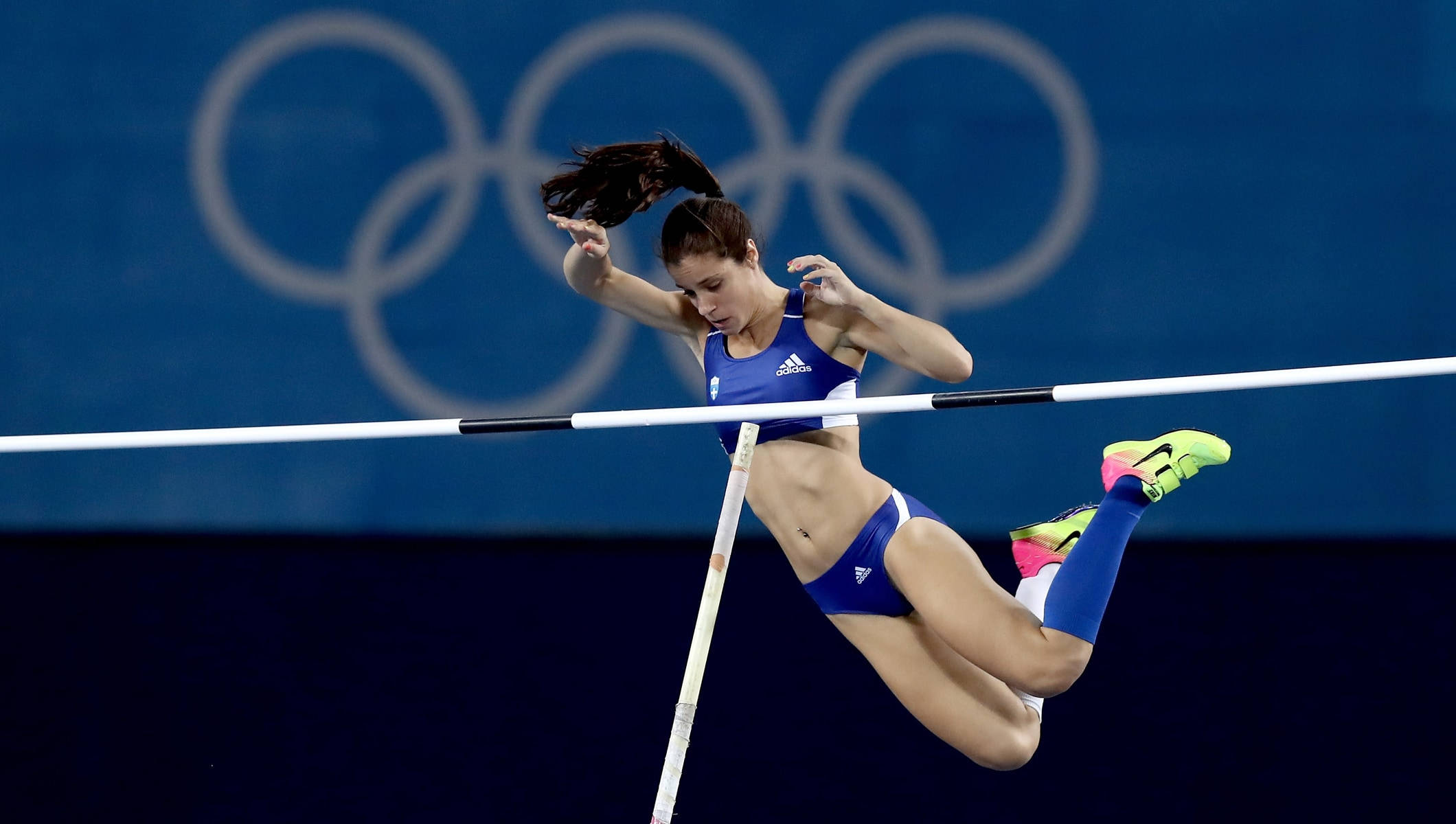 Olympics 桌布 壁紙 — 一位身穿藍色運動裝備的女子跳高選手，在藍色奧運背景下，以高度專注力越過橫桿。