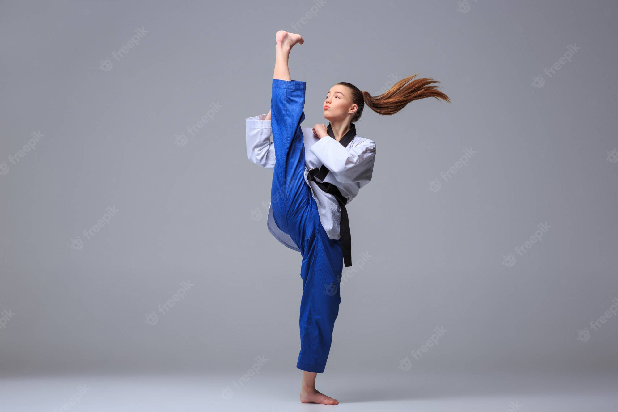 martial arts 桌布 壁紙 — 一位身穿藍色道服的女武術家，在純灰色工作室背景前，秀髮飄逸，做出高踢動作。