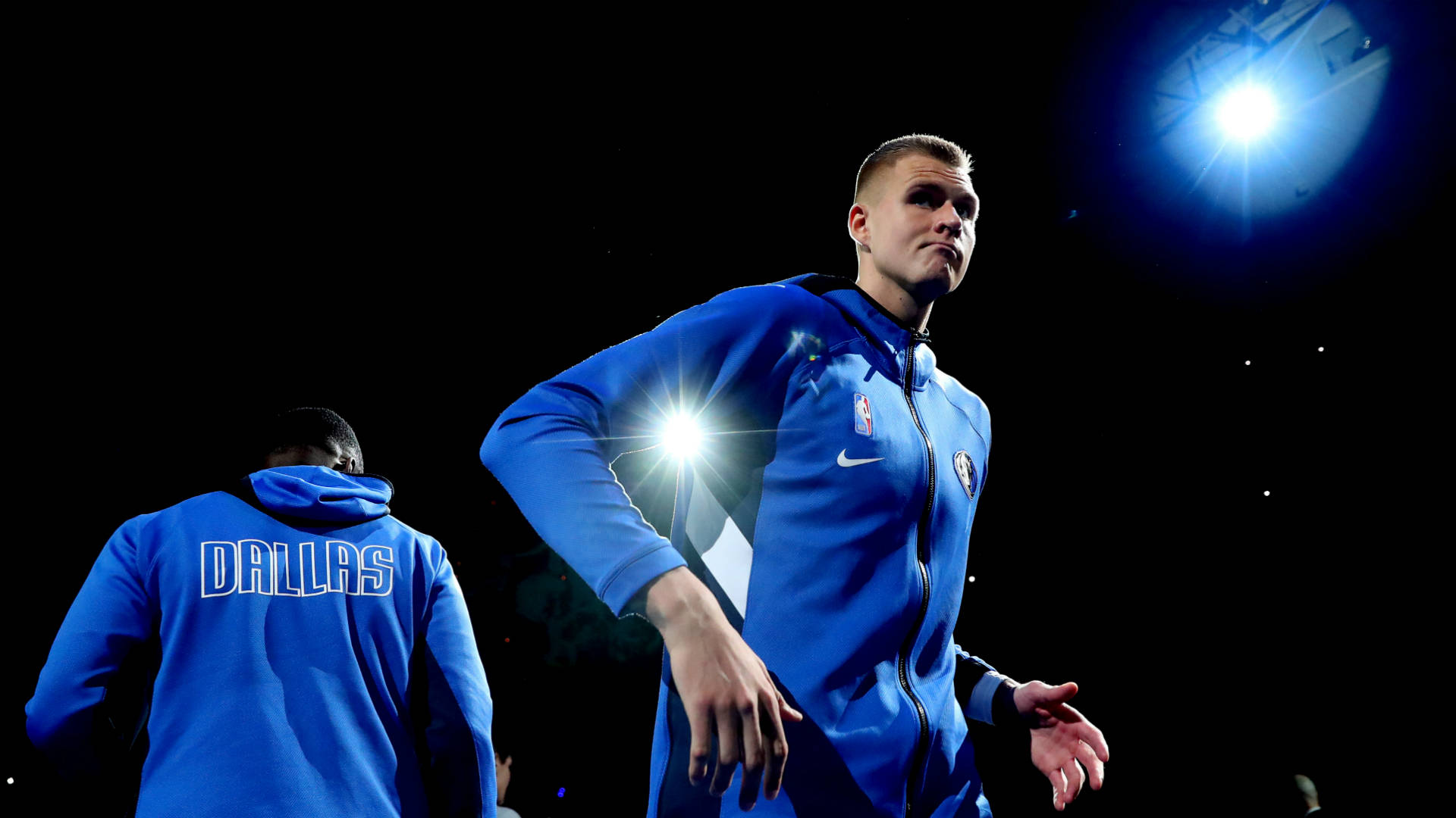 Kristaps Porzingis 桌布 壁紙 — 一位身穿藍色達拉斯連帽衫的籃球運動員站在明亮的場館燈光下，隊友正走開。