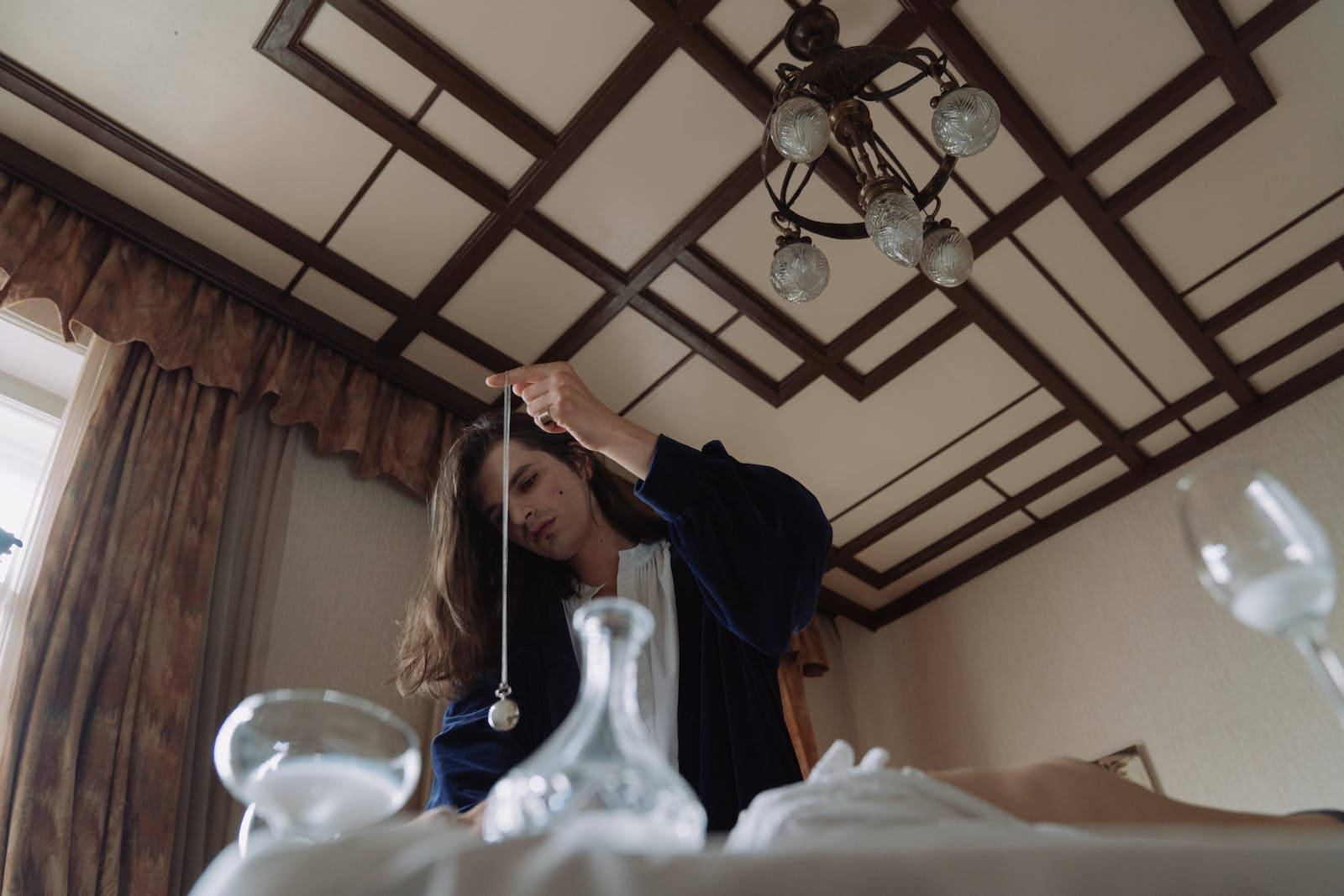 Hypnosis 桌布 壁紙 — 一位身穿藍色長袍的女性在裝飾性天花板燈下，手持鐘擺懸於擺放玻璃器皿的桌子上方。