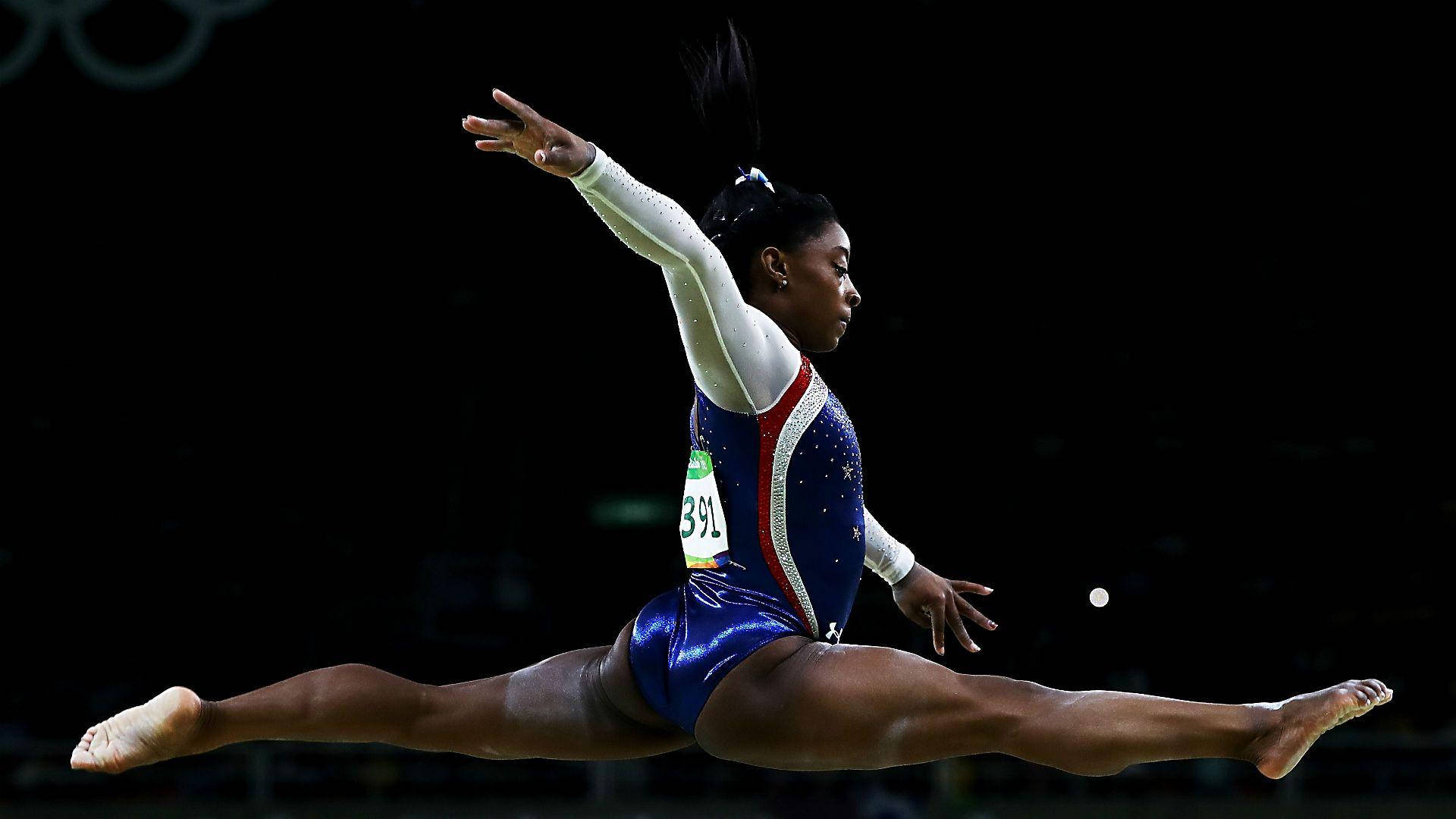 Simone Biles 桌布 壁紙 — 一位身穿藍色閃亮緊身衣的女性體操運動員在黑暗的競技場背景下進行分腿跳躍，奧運五環清晰可見。