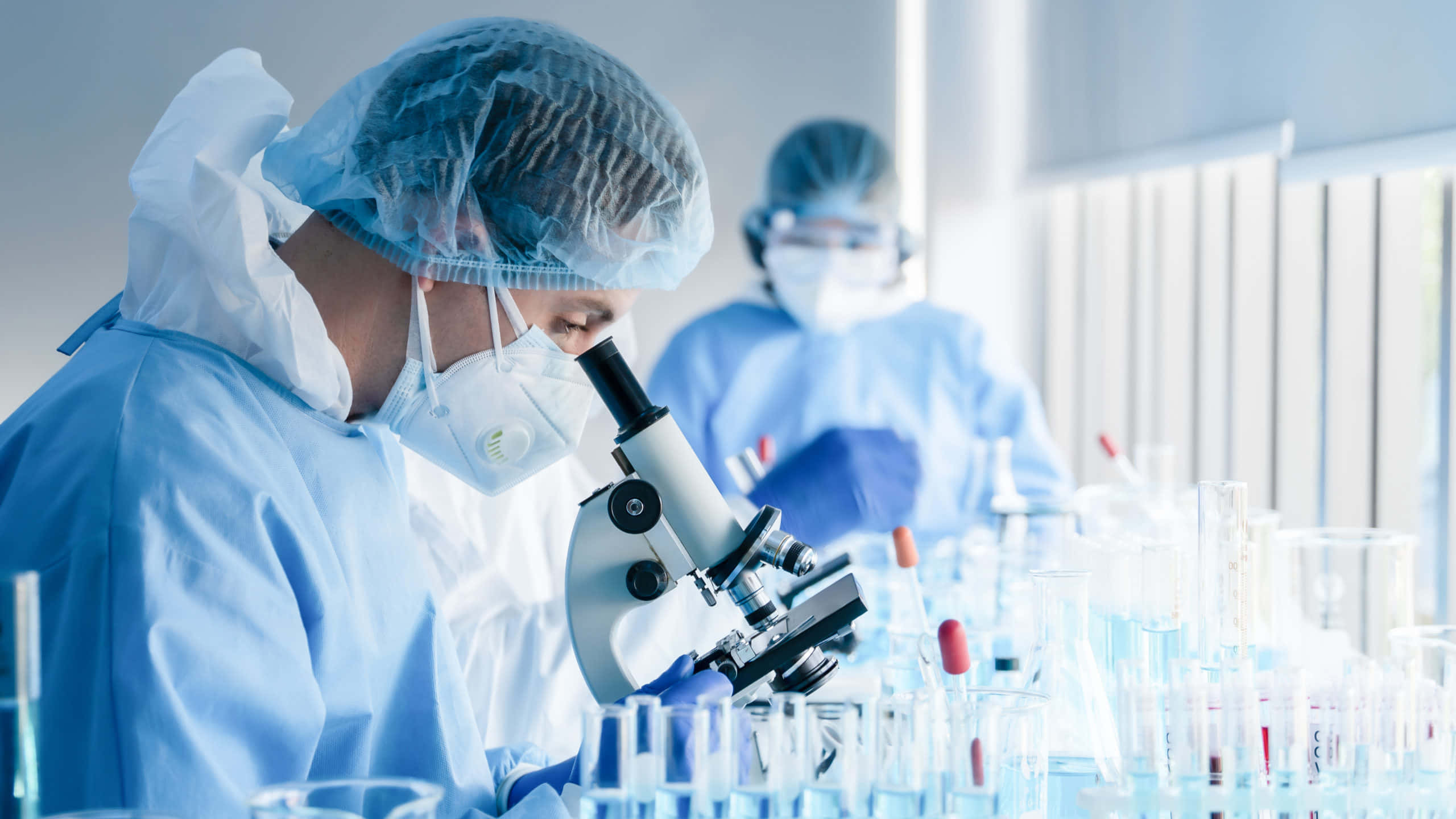科學 桌布 壁紙 — 一位身穿藍色防護裝備的科學家，在一個光線充足、裝滿玻璃器皿的實驗室裡，專注地透過顯微鏡檢查樣本。